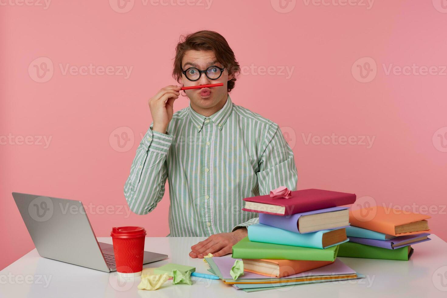 Posirive man in glasses sits by the table and working with laptop, looks at the camera, holds ipencil in lips, looks funny isolated over pink background. photo