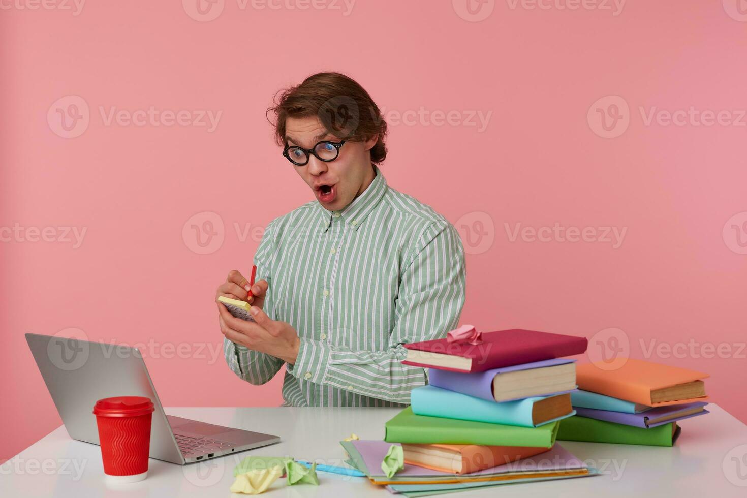 Shocked young man in glasses sits by the table and working with laptop, writes the solution of the equation on the stickers isolated over pink background. photo