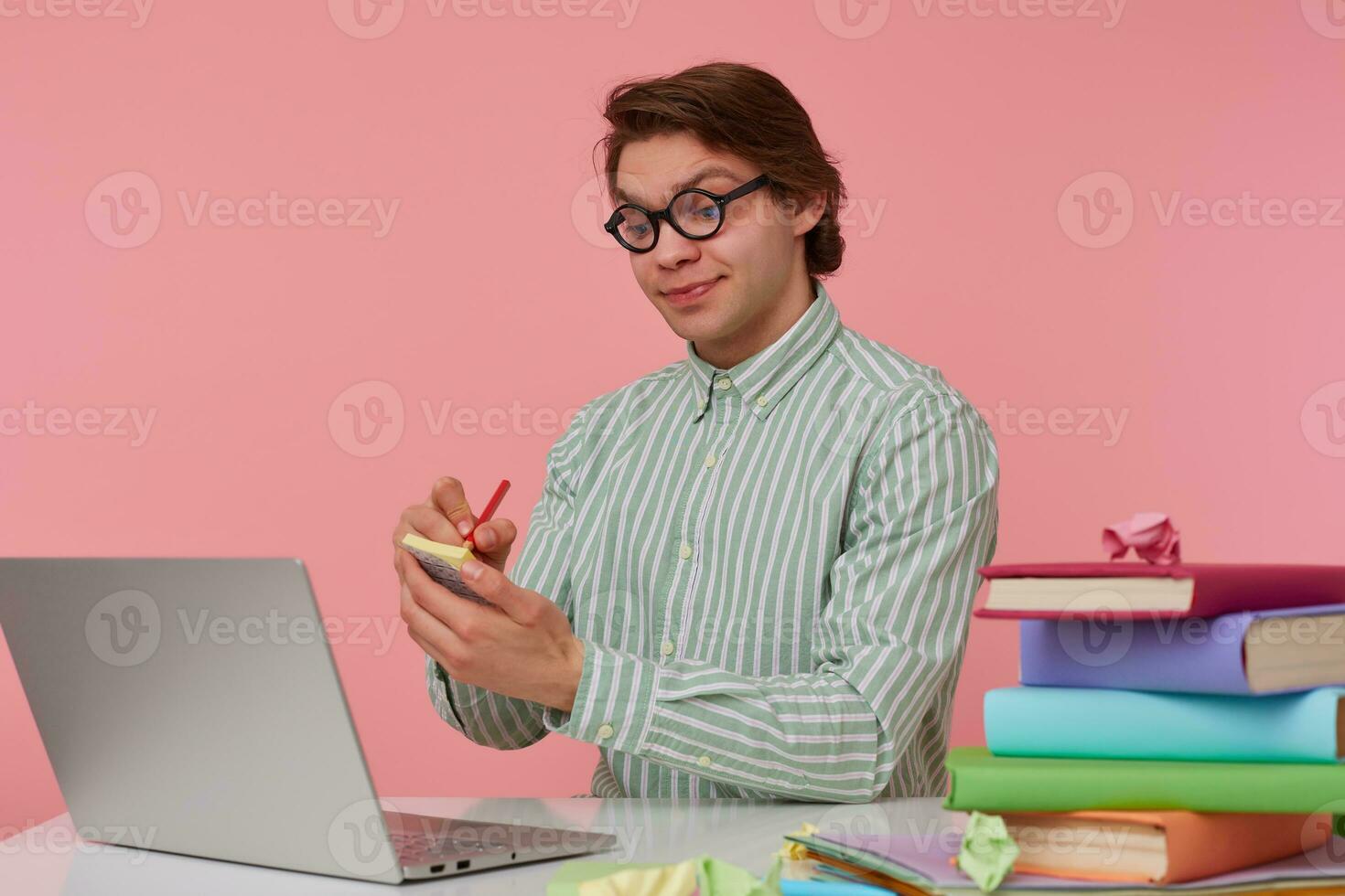 Young glad guy in glasses sits by the table and working with laptop, holds in hand a pencil, have a cool idea, pleased with himself and writes the idea on the sticker, isolated over pink background. photo