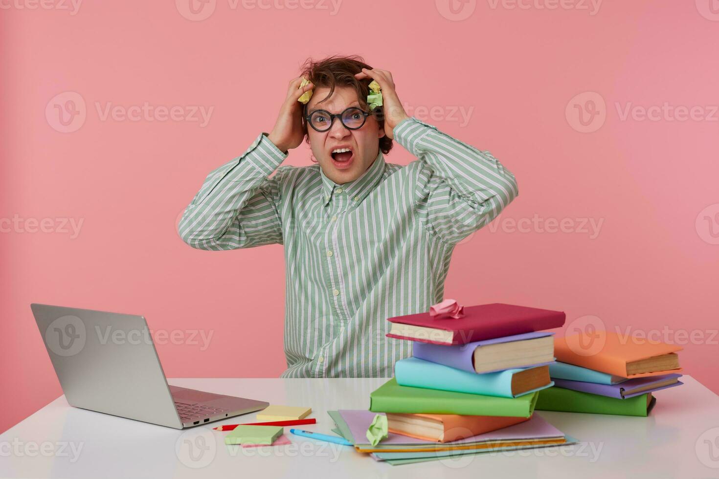 Indoor shot of stressed young dark haired male posing over pink background in striped shirt, sitting at working table with a lot of books, clutching his head with frowned face photo