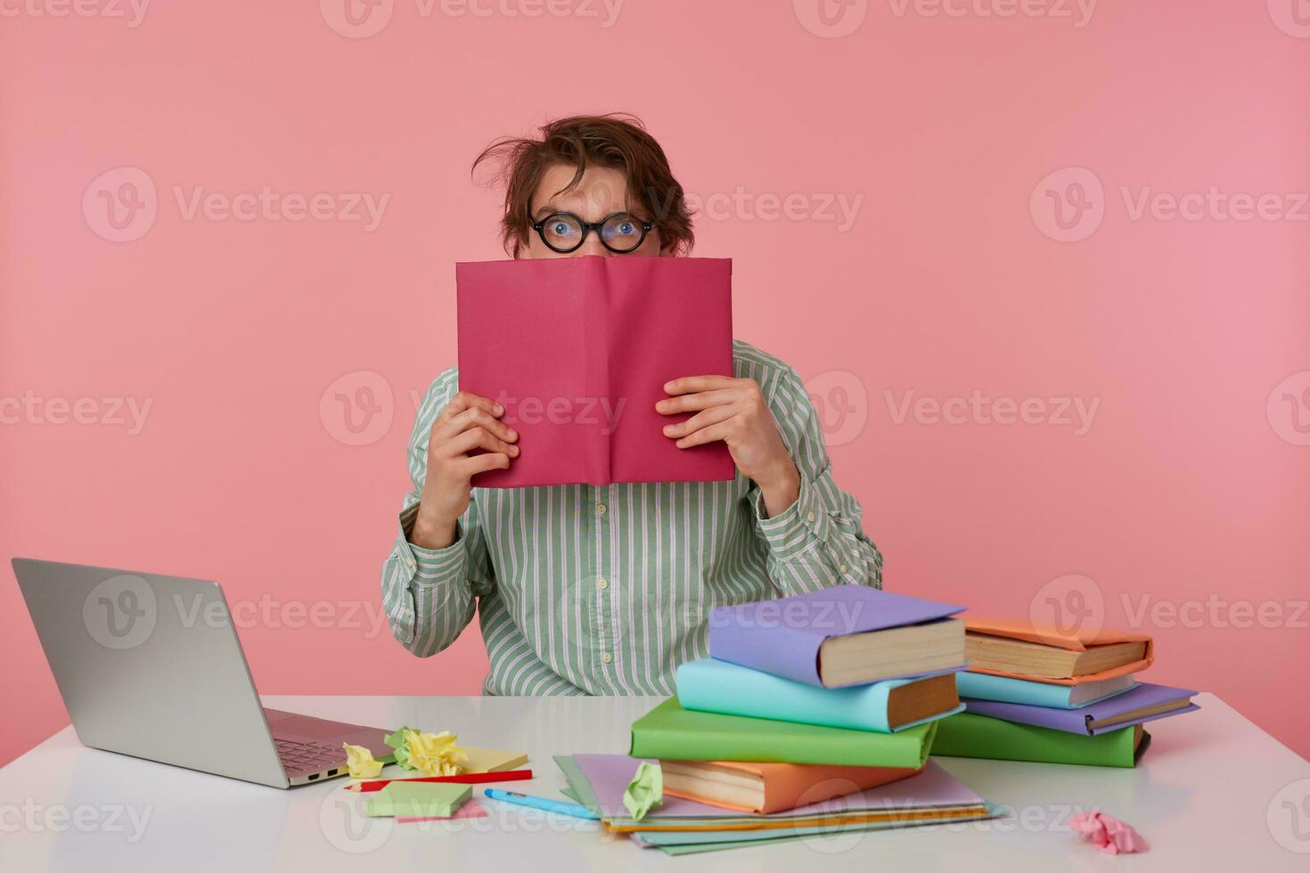 Studio photo of young dark haired male posing over pink background, wearing shirt and glasses, sitting at working table and keeping book in raised hands, looking at camera with wide eyes opened