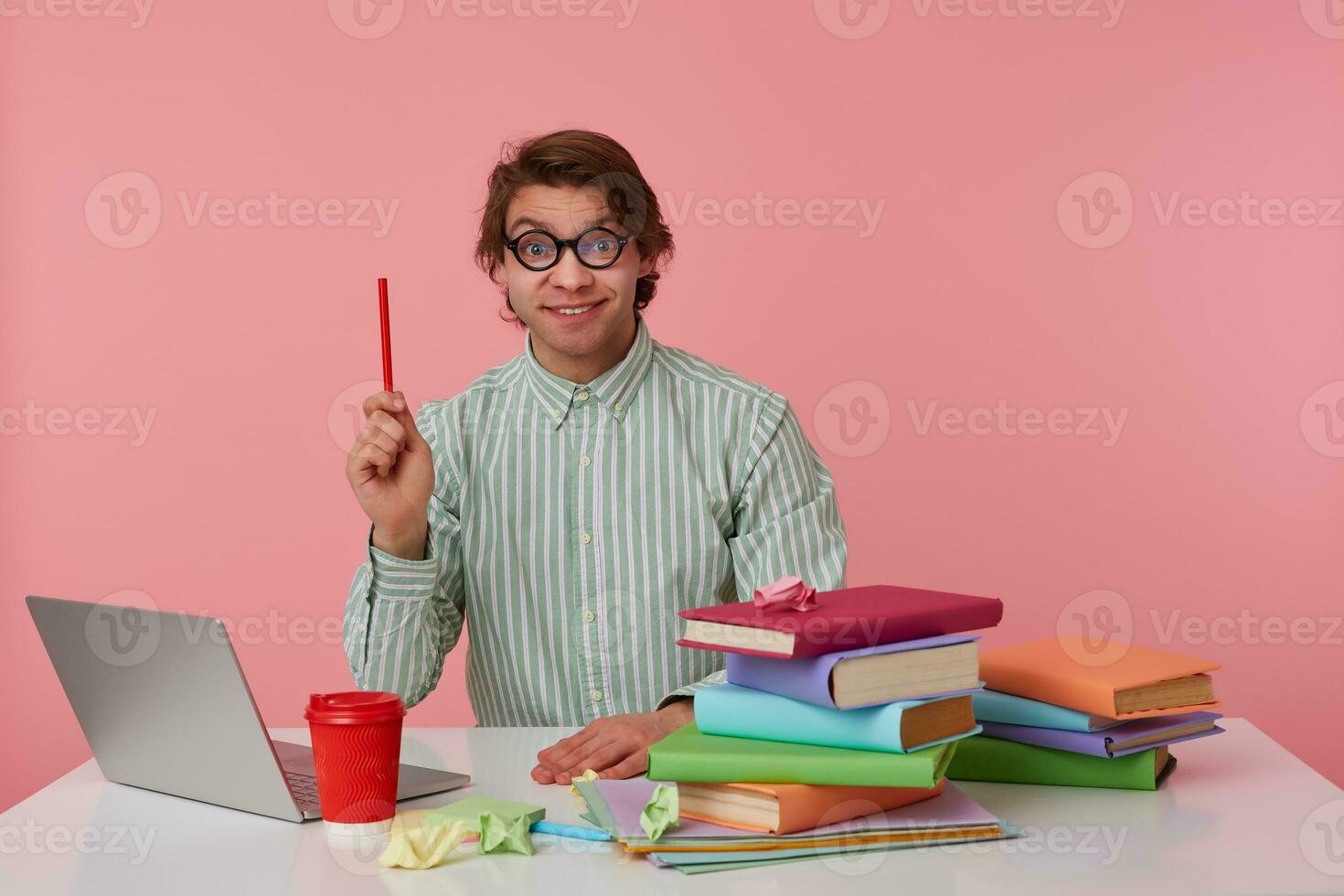 Young man in glasses sits by the table and working with laptop, looks at the camera, holds in hand a pencil, have a cool idea, isolated over pink background. photo