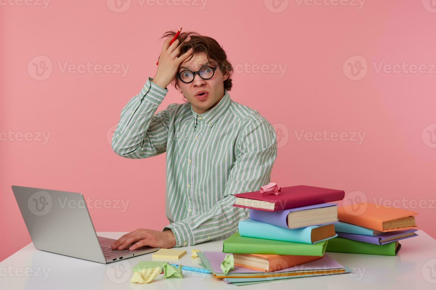 Photo of unhappy young man in glasses sits by the table and working with laptop, very dissatisfied, holds hand on forehead isolated over pink background.