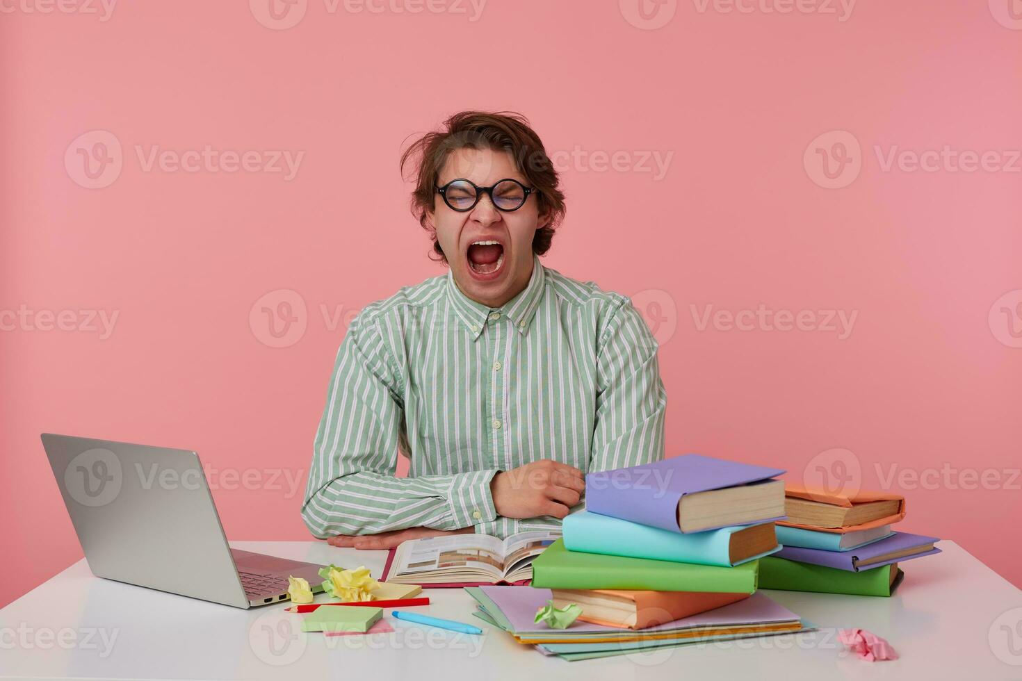 Portrait of tired young dark male with dark hair sitting at table and keeping hands on it, yawning with wide mouth opened and closed eyes, isolated over pink background photo