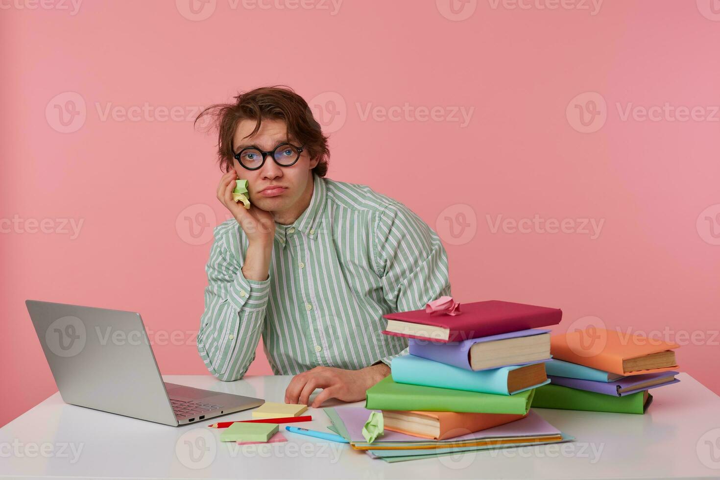 Studio photo of beat young male with wild dark hair sitting at working table over pink background, wearing shirt and eyewear, keeping chin on raised hand and looking to camera tiredly