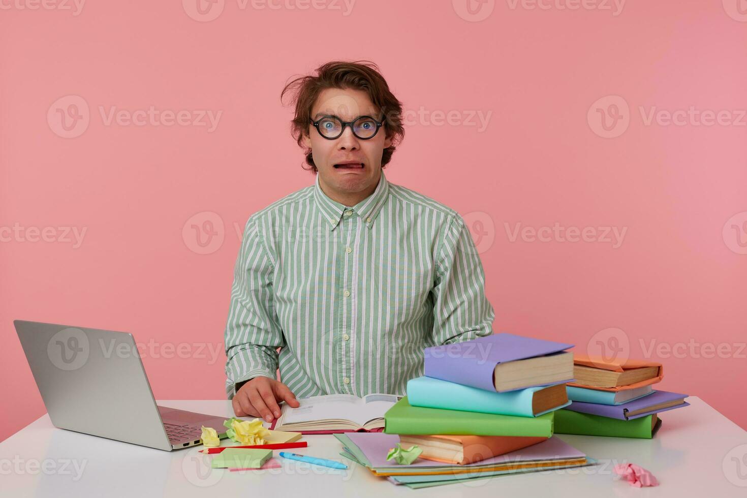 Portrait of young dark male with wild dark hair sitting at working table and making notes, grimacing over pink background in striped shirt and glasses photo