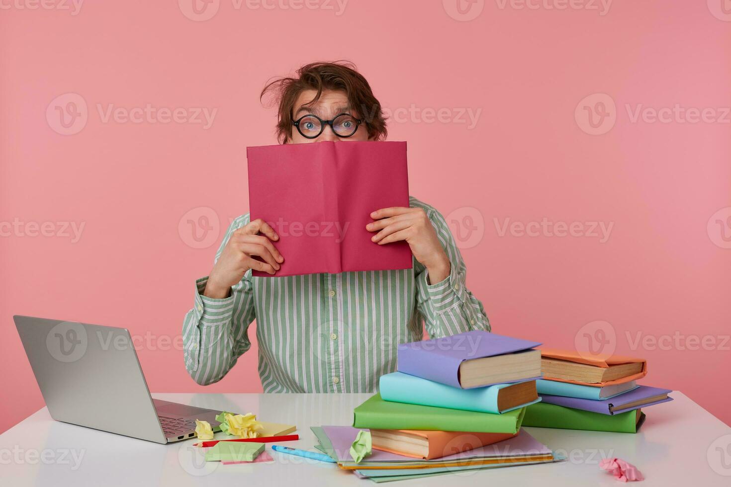 Portrait of young man with dark wild hair reading book over pink background, sitting at working with laptop and preparing for exams, wearing shirt and glasses photo