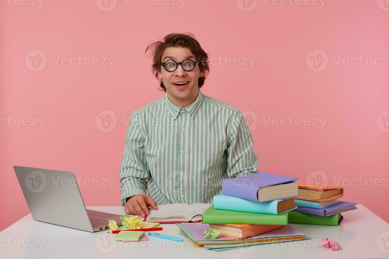 Indoor shot of happy young dark haired male in eyewear sitting at table with books and laptop, looking at camera cheerfully and raising eyebrows surprisedly, isolated over pink background photo