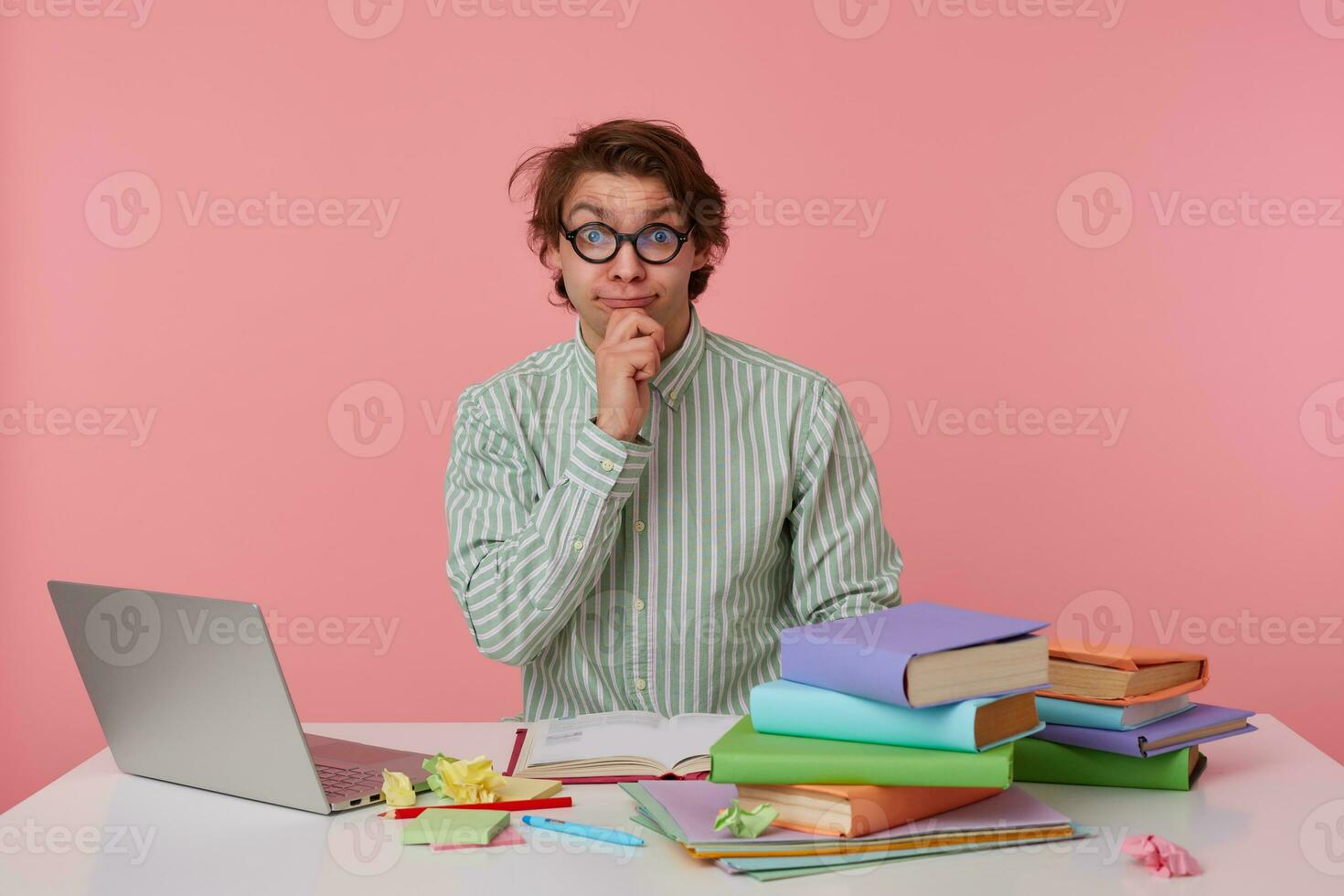 Positive young male with wild hair sitting at working table with books and laptop, posing over pink background in shirt and glasses, holding chin with hand and looking to camera with raised eyebrows photo