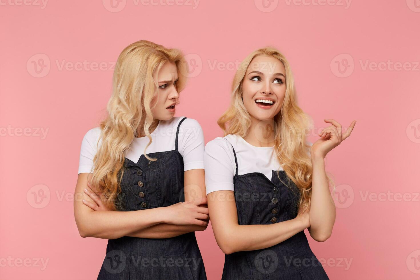 perplejo joven largo peludo rubia hembra cruce manos en su cofre mientras mirando confusamente en su de cabeza blanca alegre hermana, en pie terminado rosado antecedentes foto