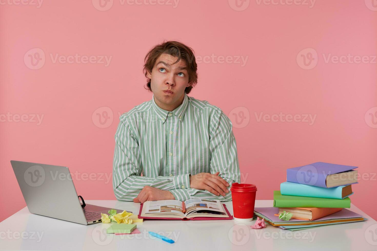 Indoor shot of thoughtful young dark haired male posing over pink background, sitting at working table with folded arms on it, looking upwards with pursed lips and raising eyebrows photo