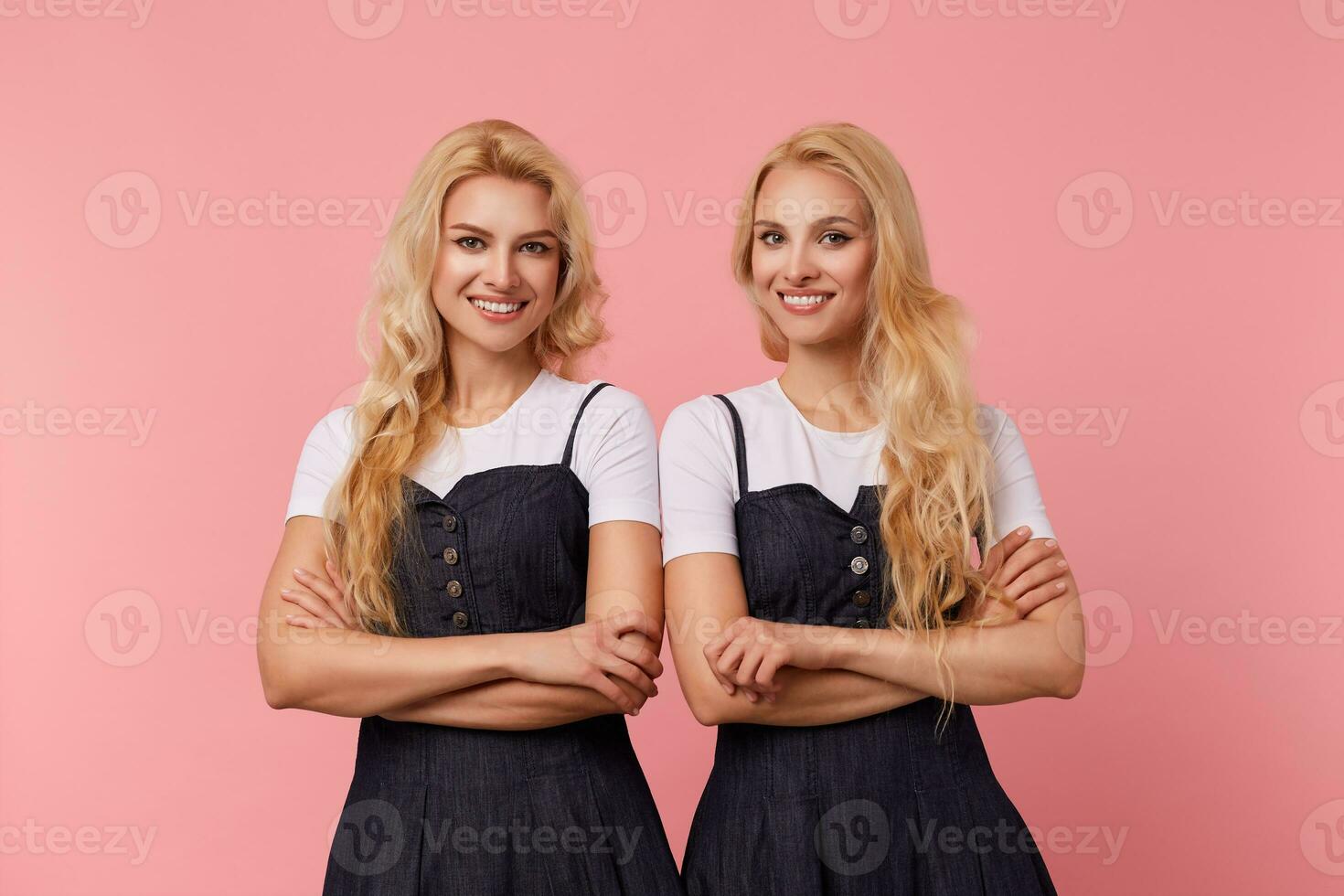 Indoor photo of young cheerful long haired blonde females keeping hands crossed while looking gladly at camera with wide smiles, standing over pink background