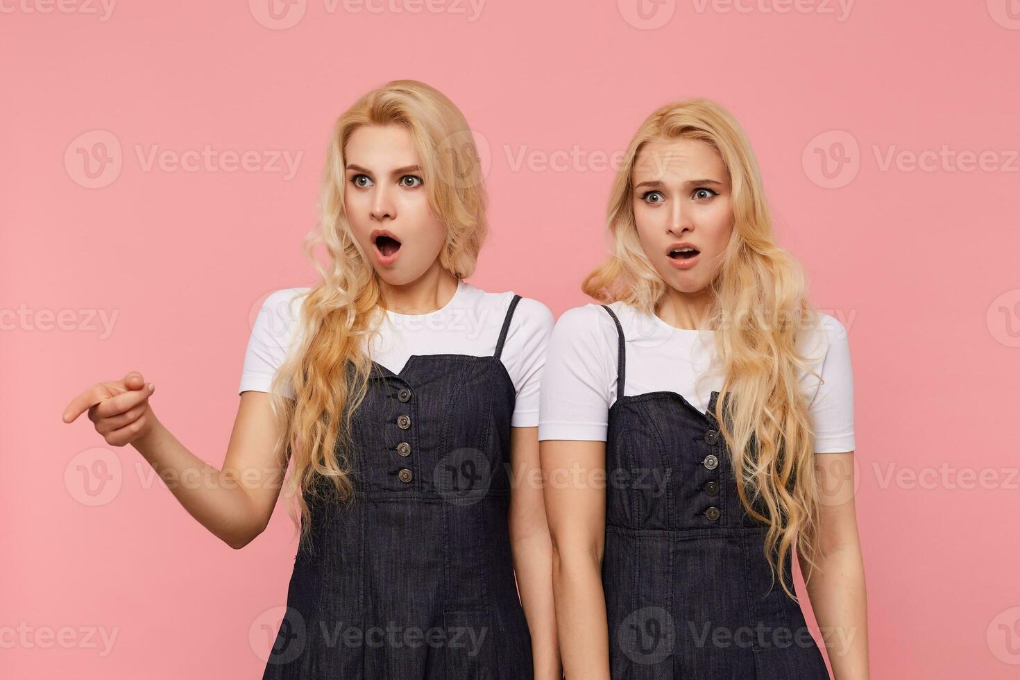 Horizontal shot of young bewildered long haired white-headed females keeping their mouths opened while looking confusedly aside, isolated over pink background photo