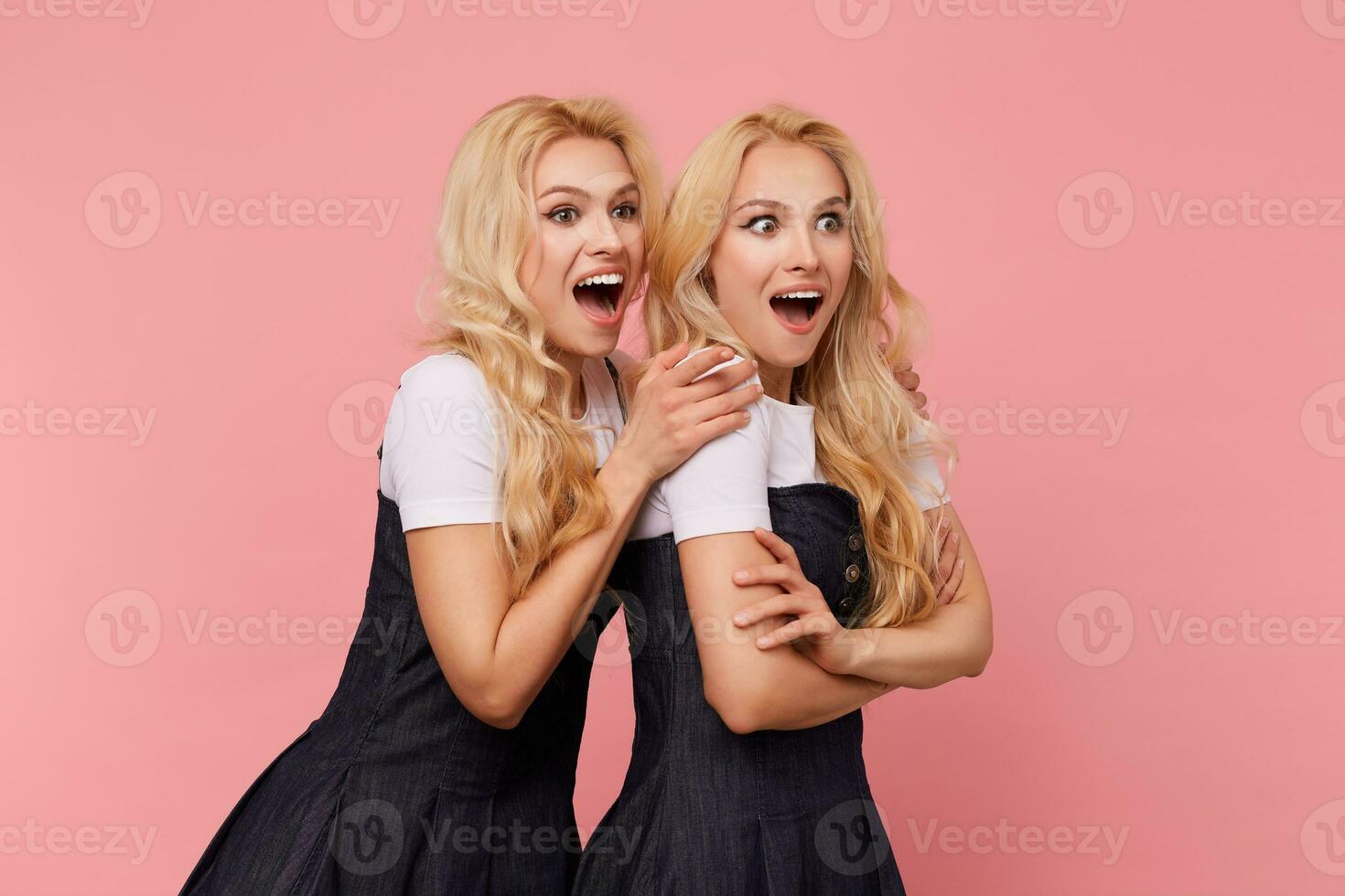 Excited young beautiful long haired blonde women looking surprisedly aside with wide mouths opened while standing over pink background in elegant clothes photo