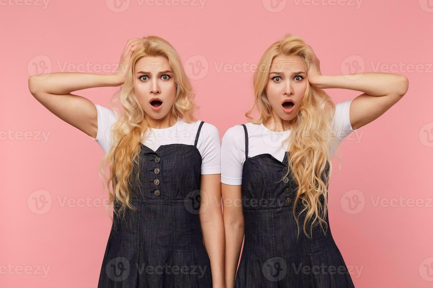 Shocked young lovely long haired blonde women keeping raised palms on their heads while looking dazedly at camera with wide mouths opened, isolated over pink background photo