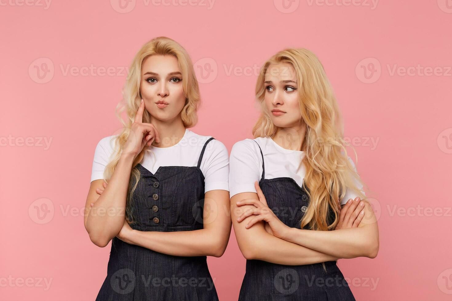 Indoor photo of young brown-eyed long haired female keeping forefinger raised while looking attentively at camera, posing over pink background with her confused pretty blonde sister