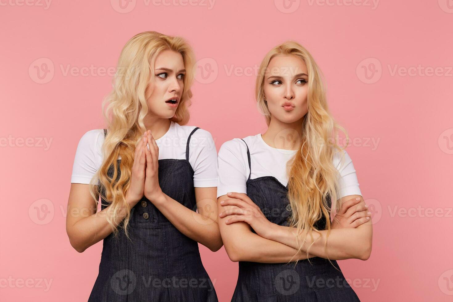 Studio shot of young pretty bewildered long haired lady keeping raised palms folded while looking surprisedly on her pensive pretty white-headed sister, isolated over pink background photo