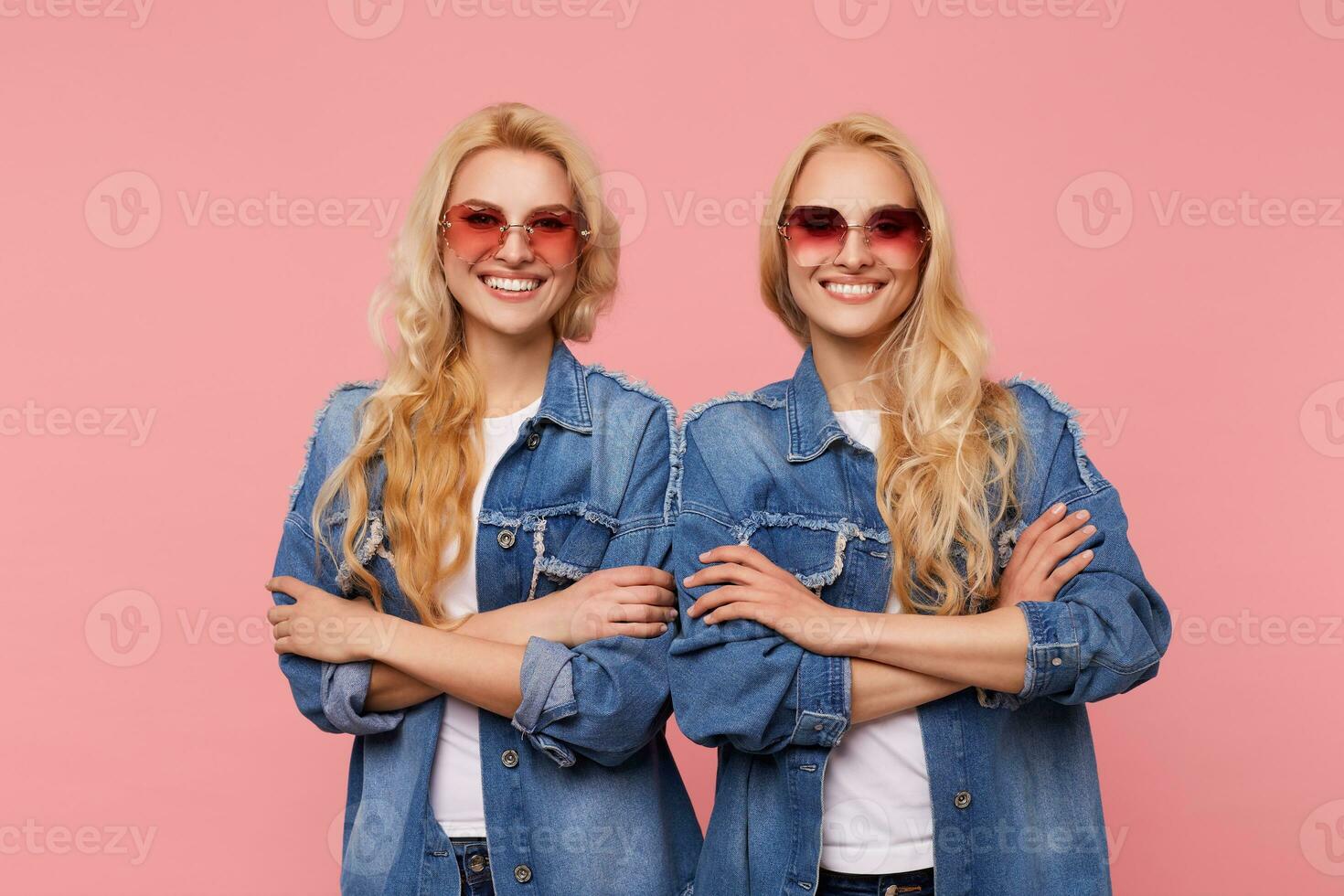 Two beautiful white-headed females in sunglasses and jeans coats folding their hands on chest while standing over pink background, smiling widely while looking cheerfully at camera photo