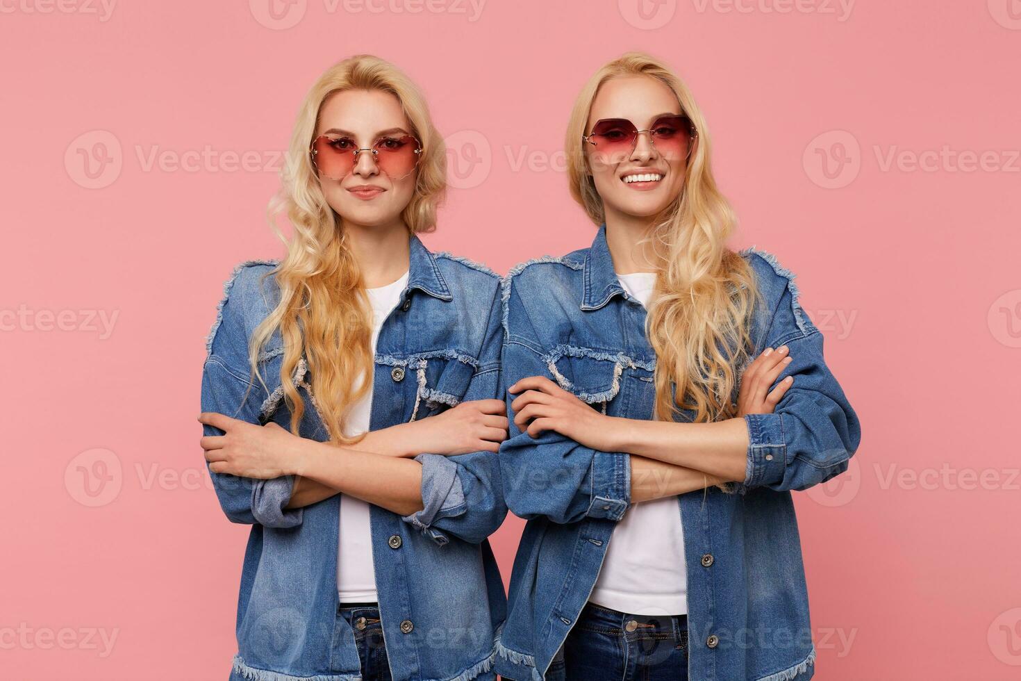 Positive young attractive long haired blonde women in red sunglasses looking at camera with pleasant smiles and keeping hands crossed while standing over pink background photo