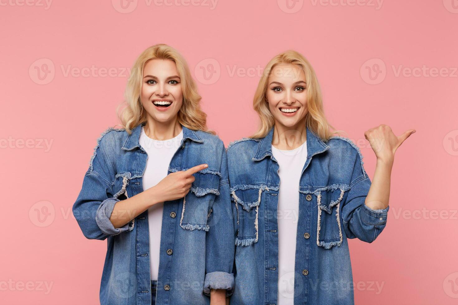 Happy young attractive long haired blonde ladies showing their perfect white teeth while smiling cheerfully and thumbing aside, isolated over pink background photo