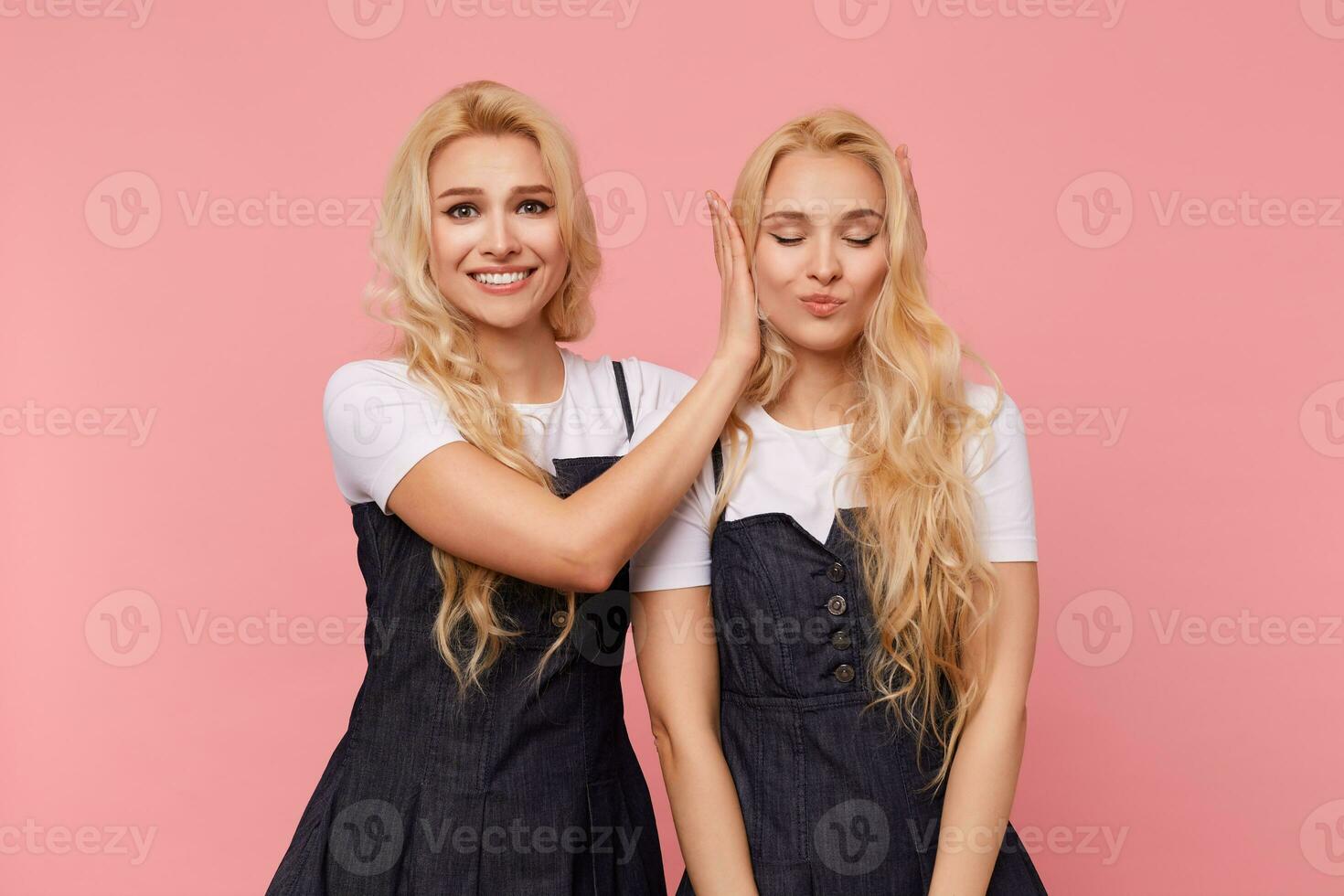 Happy young brown-eyed long haired female smiling widely while looking at camera and covering ears of her pretty blonde sister while making her surprise, isolated over pink background photo