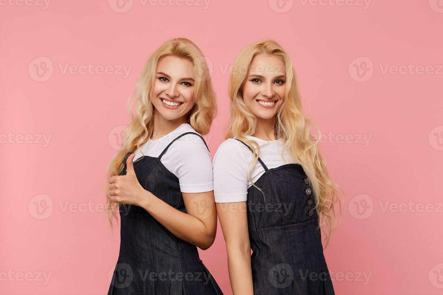 Studio photo of young long haired white-headed ladies showing their white perfect teeth while looking happily at camera with wide smiles, isolated over pink background