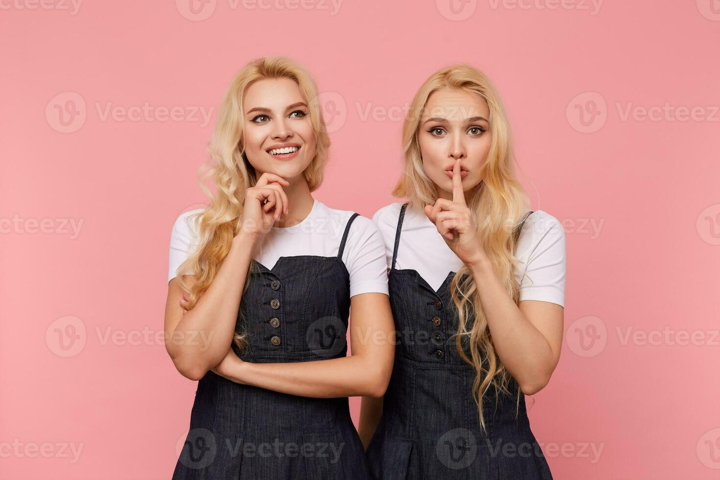 Cheerful lovely white-headed lady with loose hair holding her chin with raised hand and smiling happily while her blonde serious sister raising hand with hush gesture to her mouth photo