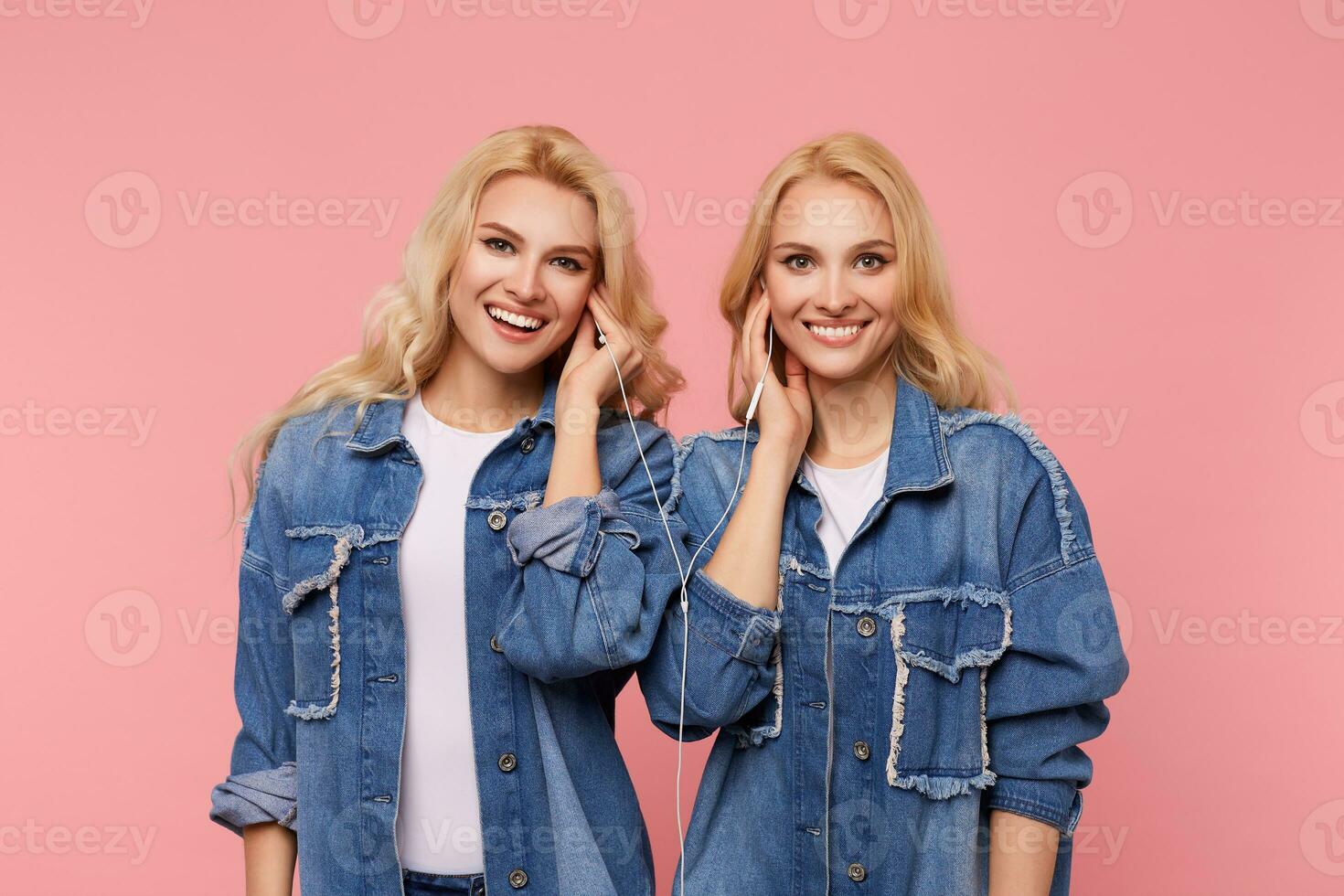 Happy young attractive long haired blonde sisters looking at camera with charming smiles while listening to music in headphones, isolated over pink background photo