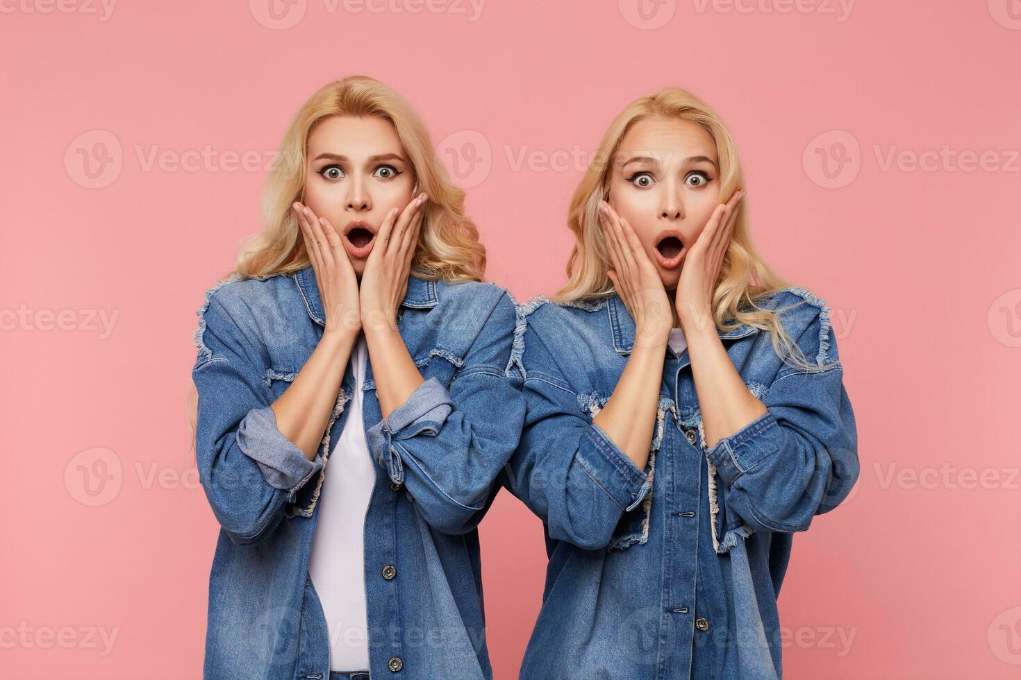 Shocked young pretty white-headed sisters with curls holding their faces with raised palms while looking dazedly at camera with wide eyes opened, isolated over pink background photo