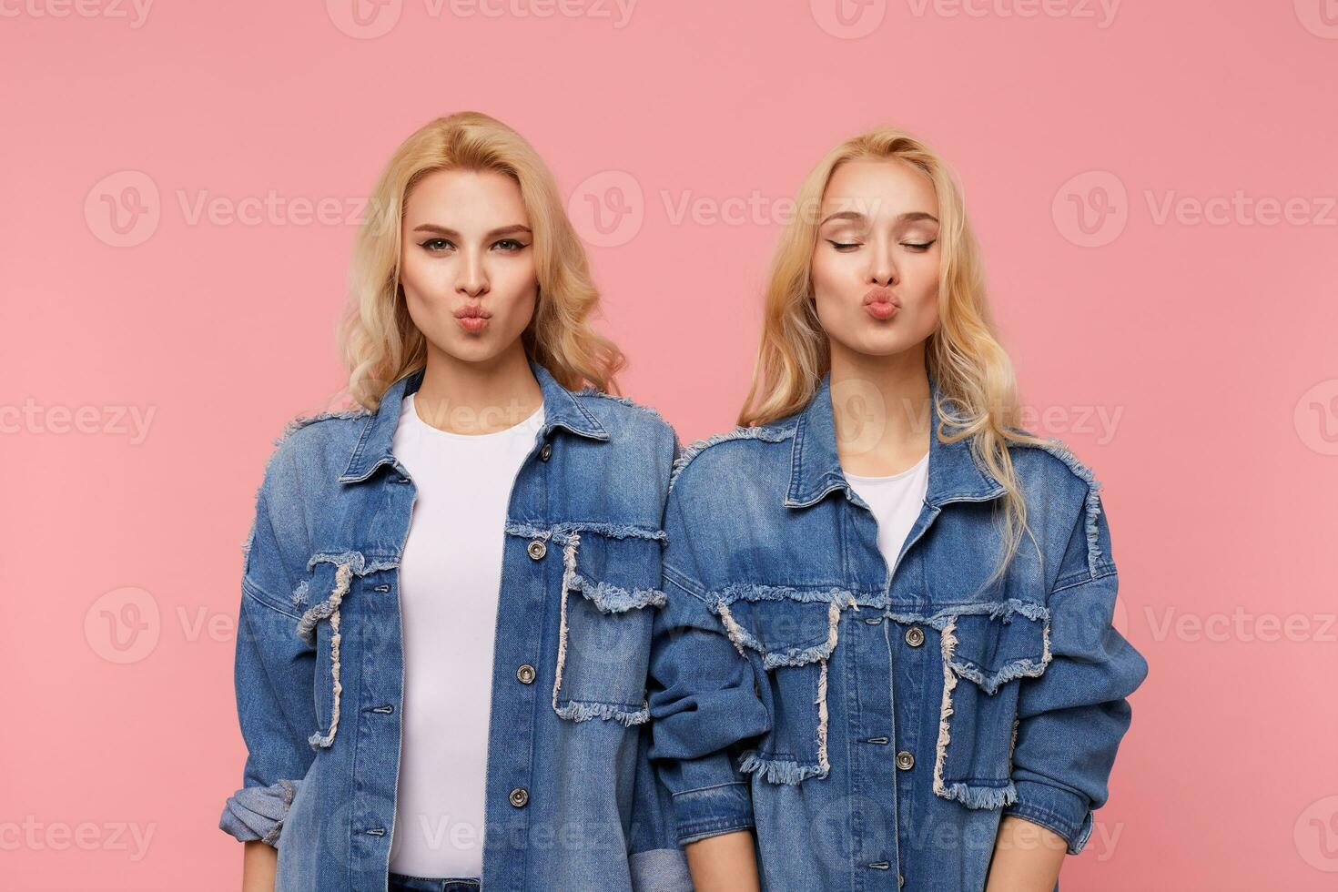 Posititve attractive young blonde sisters with curls folding lips in air kiss while standing against pink background and keeping hands along their bodies photo