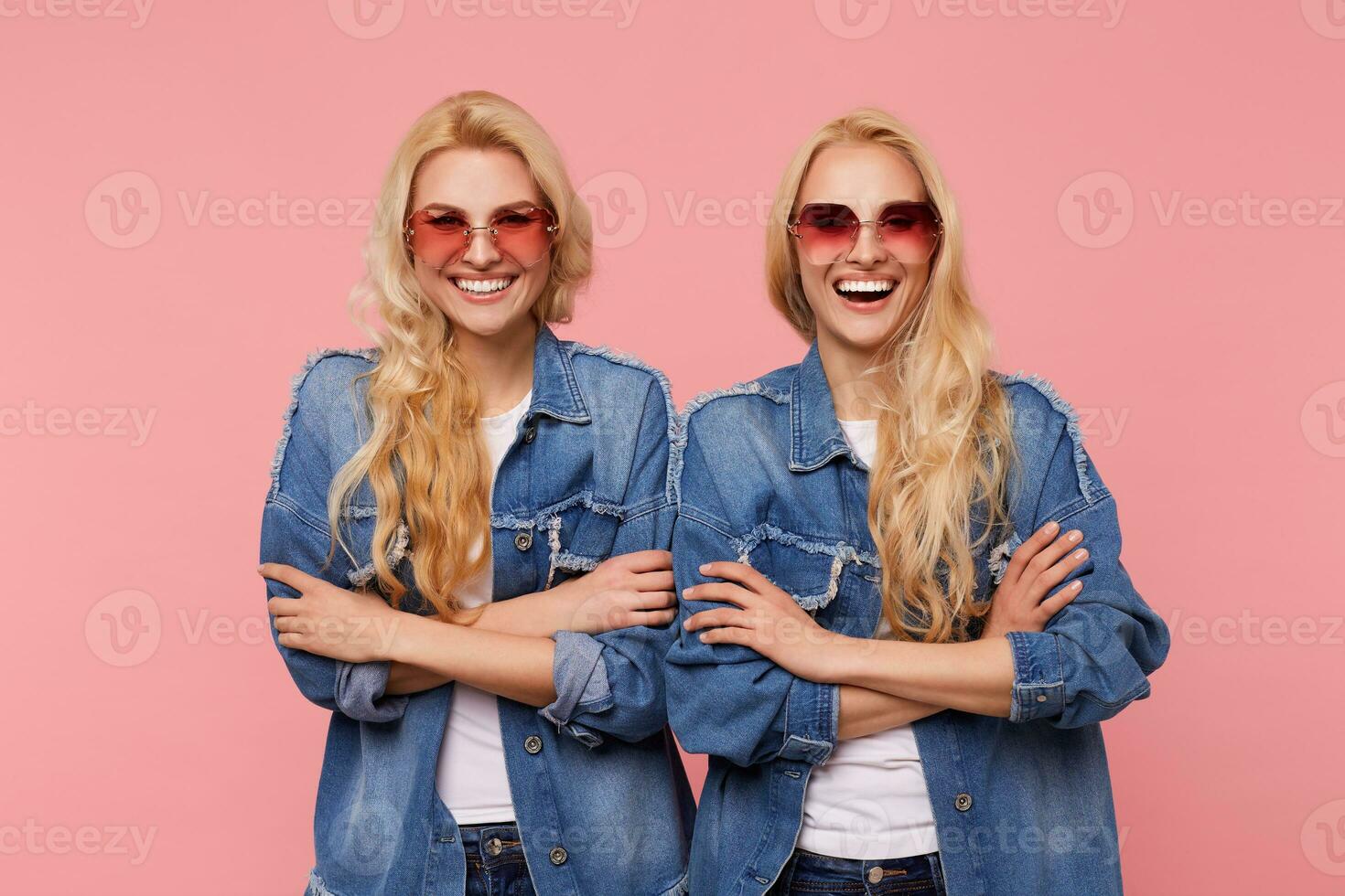 Joyful young lovely long haired blonde twins keeping hands crossed while looking happily at camera with broad smiles, isolated against pink background photo