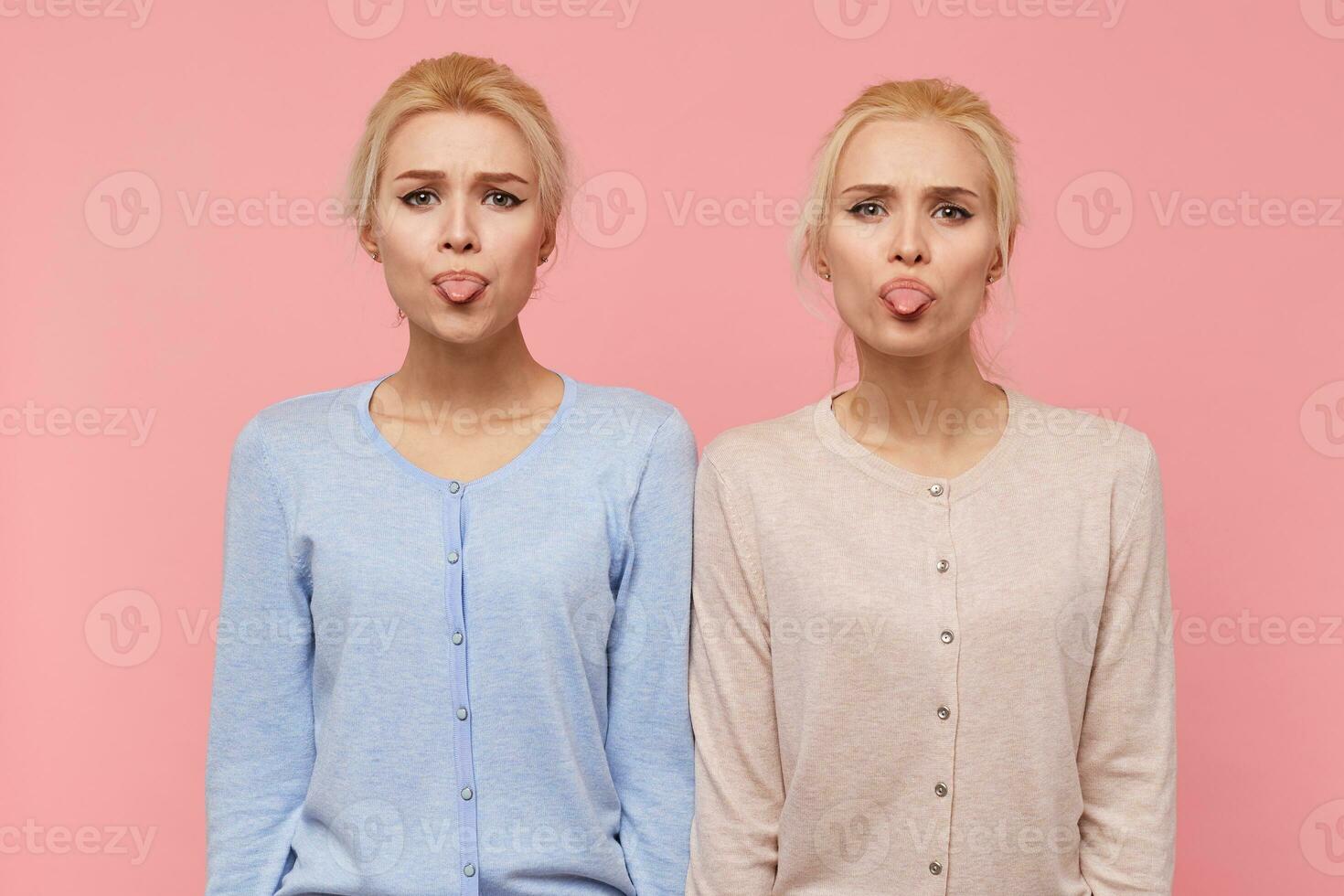 Portrait of beautiful young blonde twins make faces and show tongues, looking at the camera isolated over pink background. photo