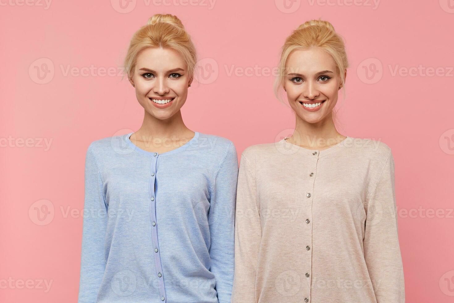 Beautiful young blonde twins in a good mood, broadly smiling in the camera isolated over pink background. photo