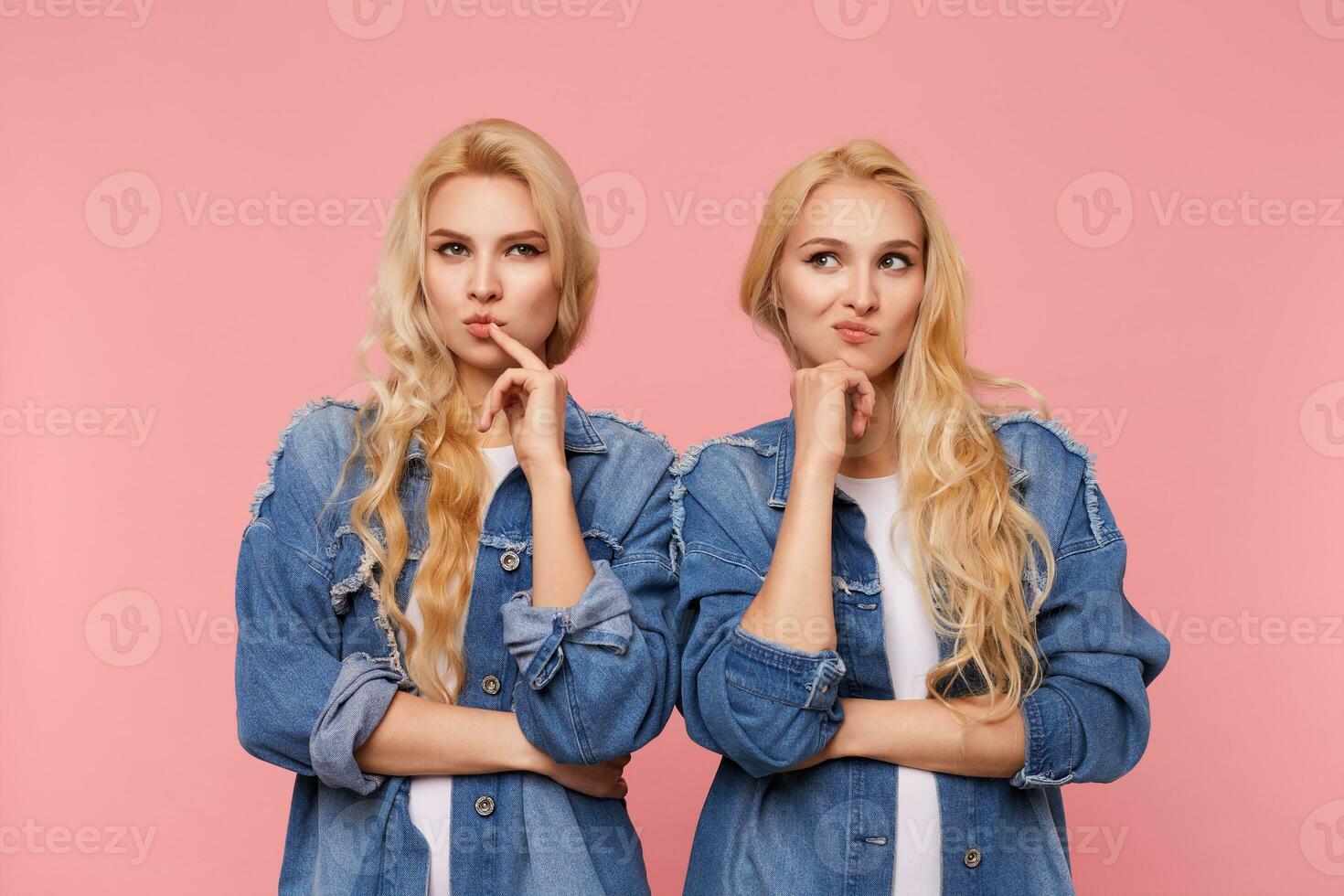 Puzzled young attractive twins curly hairstyle touching face with raised hands and twisting pensively mouth while standing over pink background in jeans coats photo