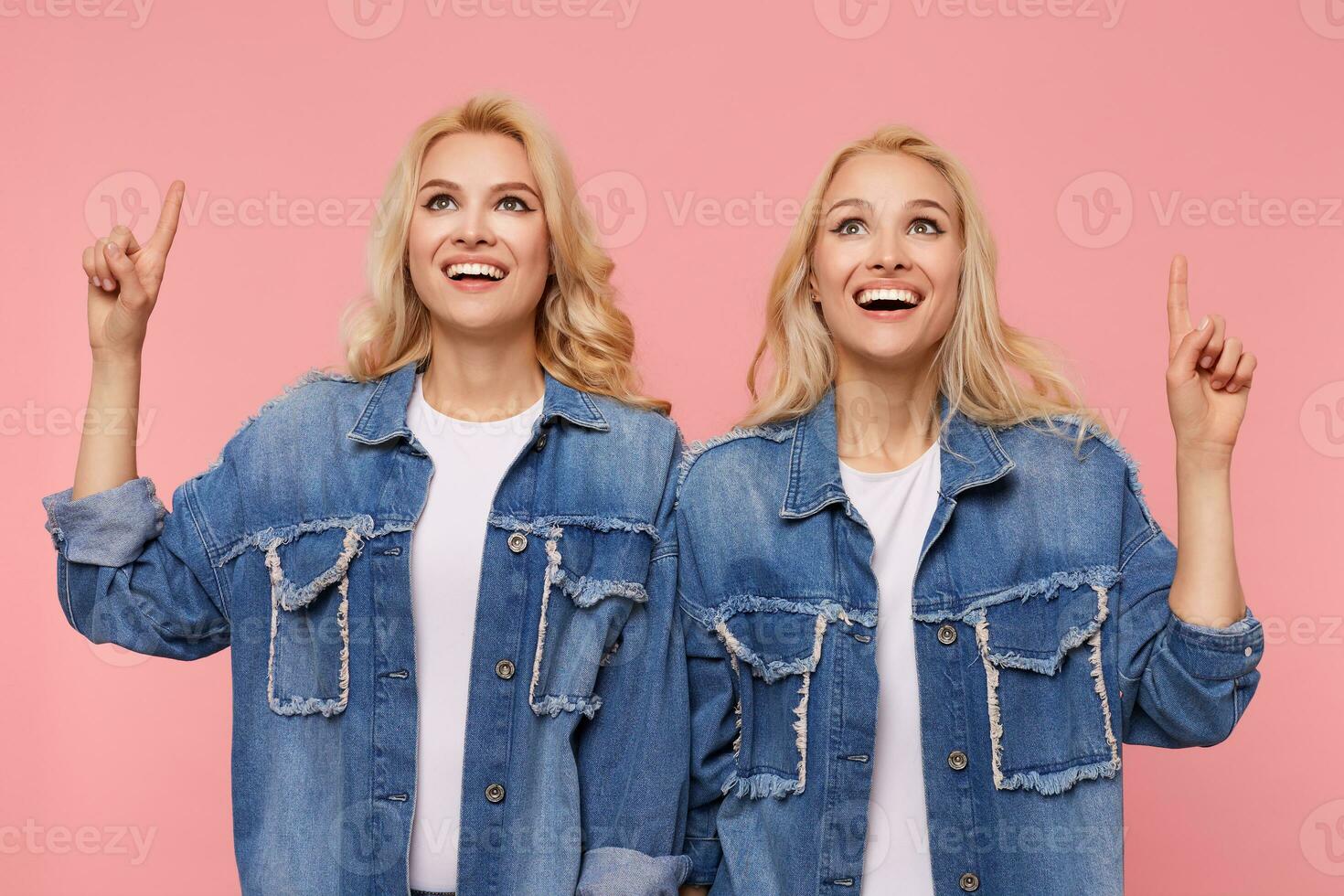 Indoor shot of young joyful long haired blonde twins smiling cheerfully while pointing upwards with forefingers, standing over pink background in casual clothes photo