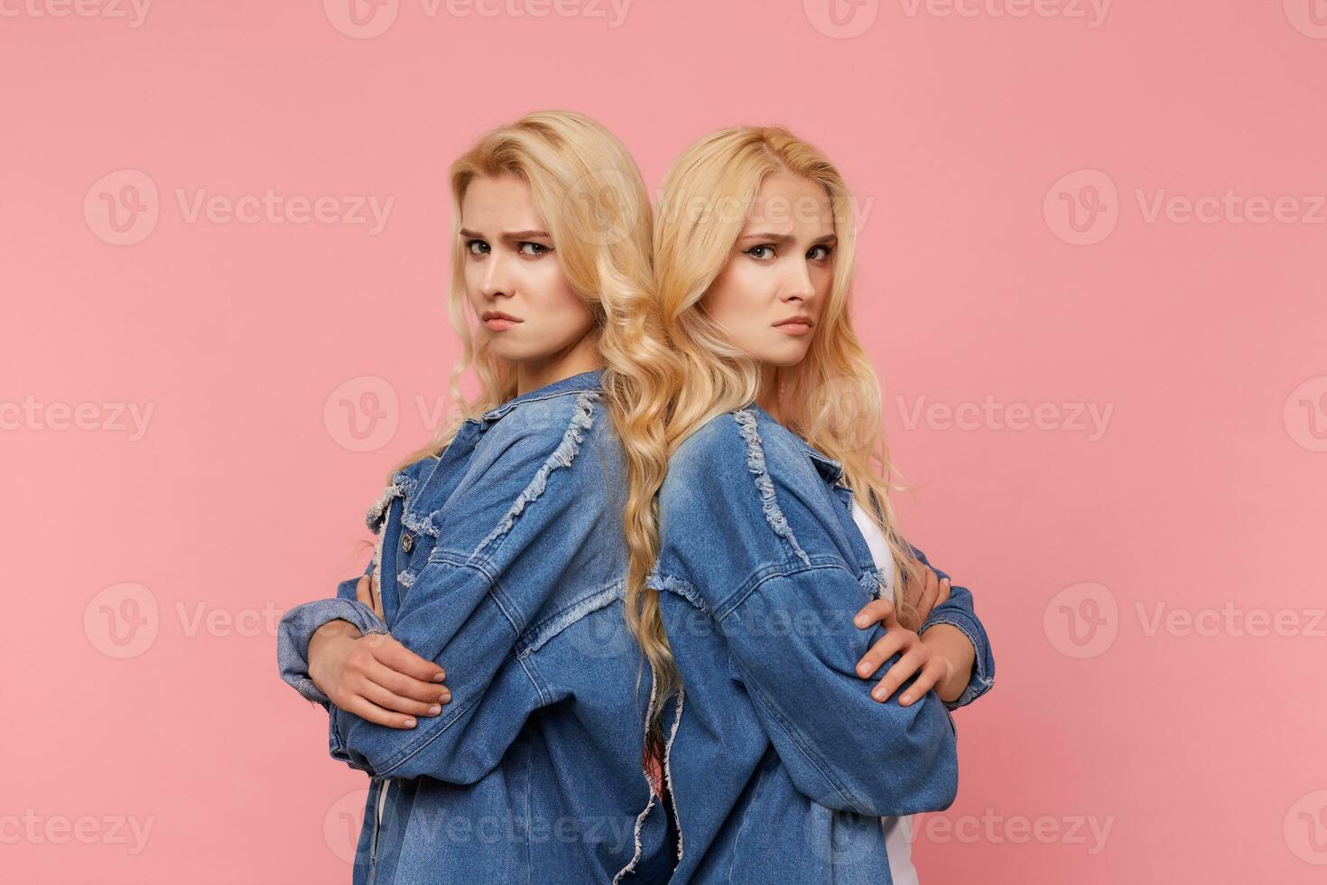 Unhappy young pretty long haired blonde sisters folding hands on chests while looking severely at camera, standing against pink background back to back photo