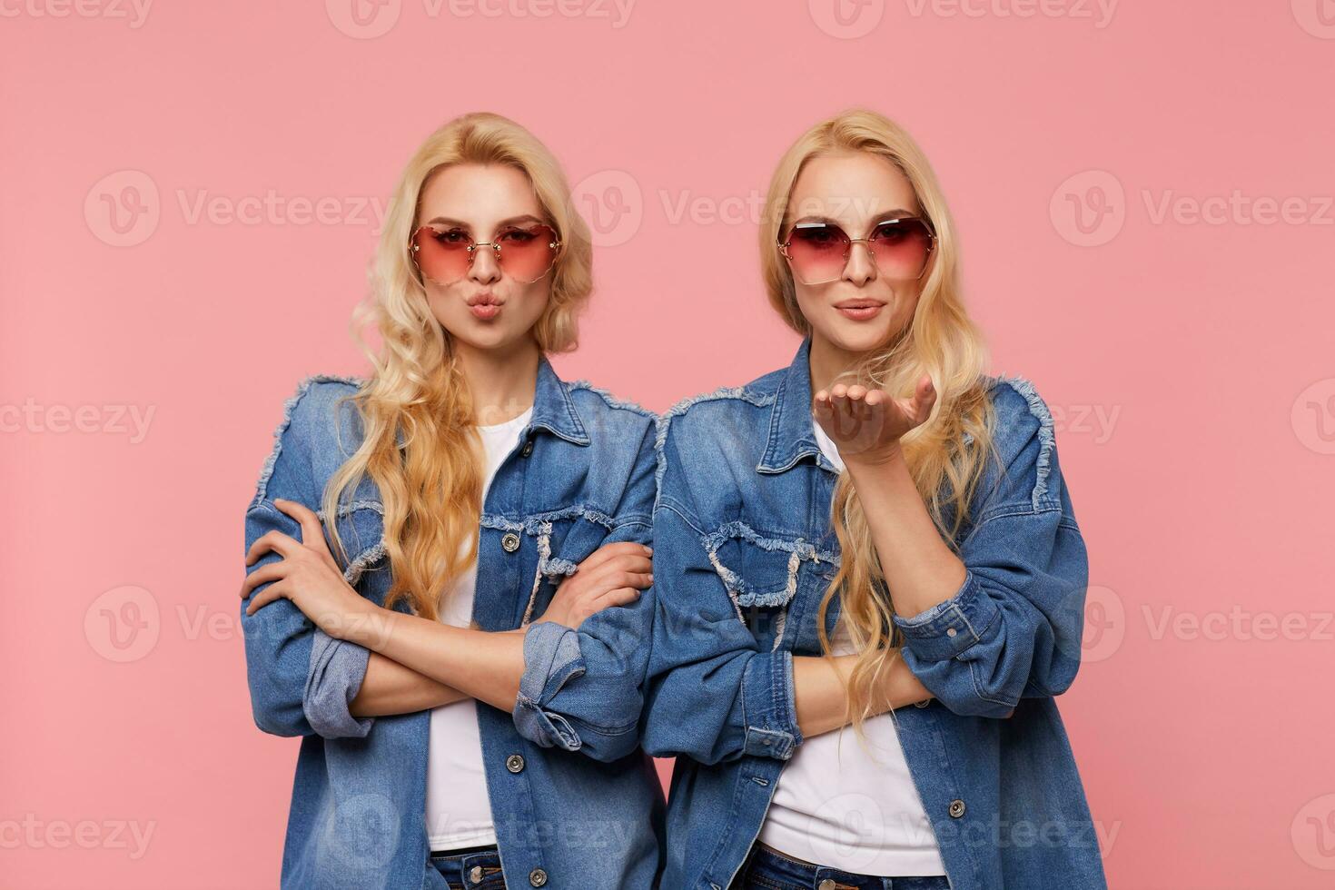 Horizontal photo of young beautiful long haired females with wavy hairstyle folding lips in air kiss while looking positively at camera, isolated over pink background