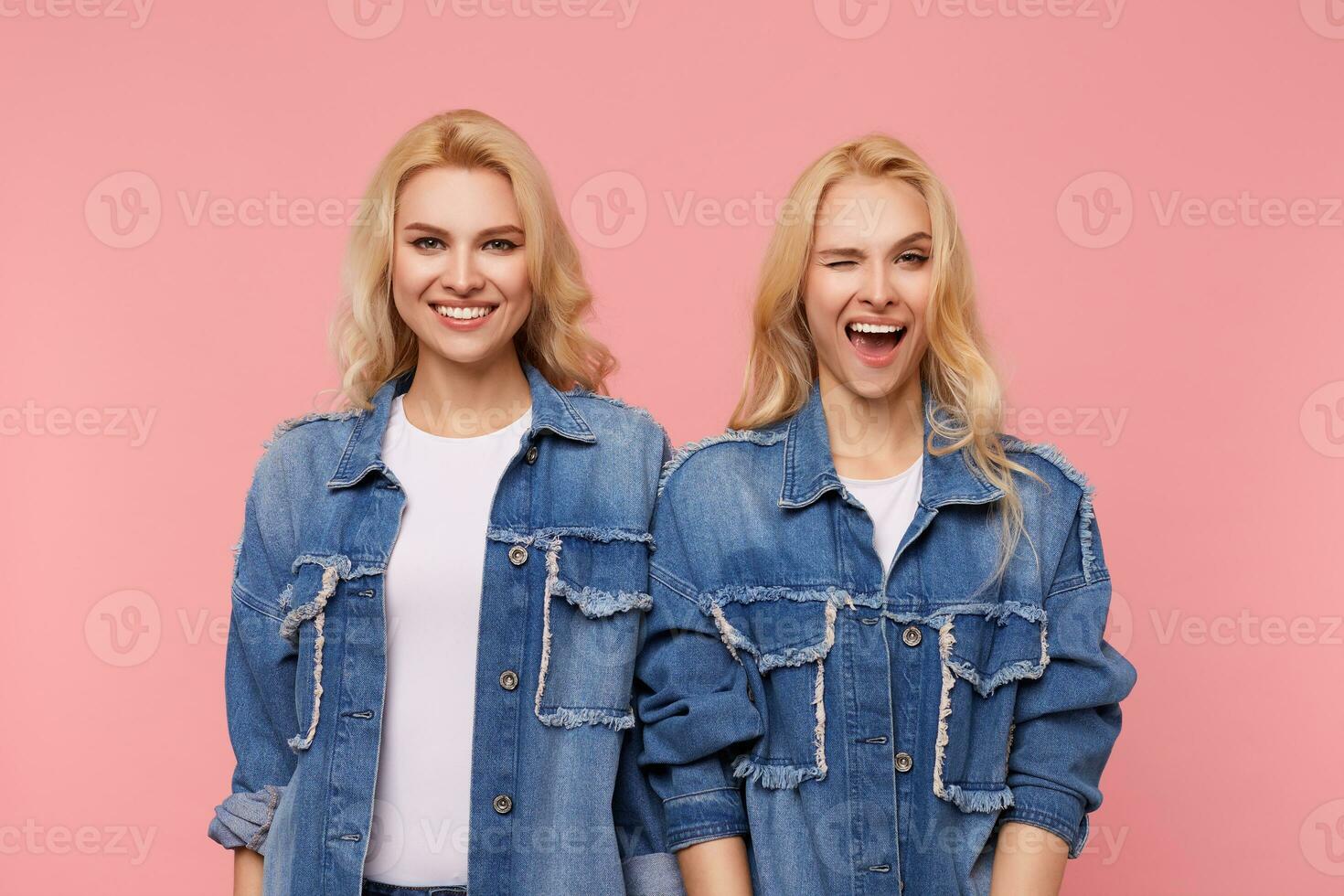 Studio photo of young lovely fair-haired women with wavy hairstyle being in nice mood and smiling hapily while standing over pink background in casual clothes