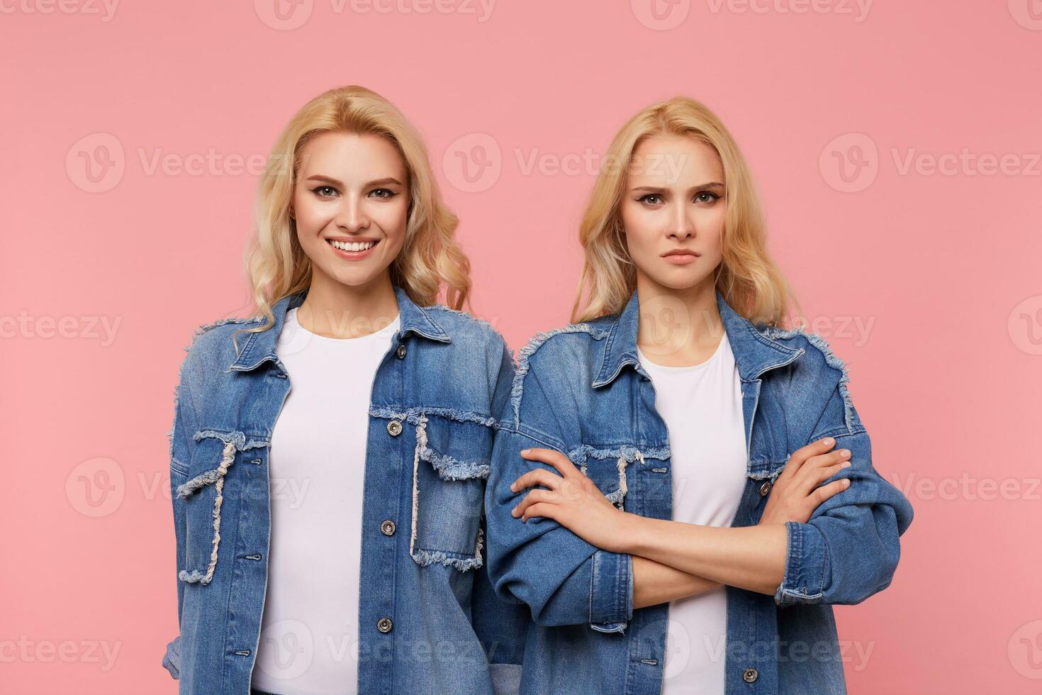 Indoor shot of young severe blonde lady keeping hands folded while standing over pink background with cheerful blue-eyed pretty blonde woman with wavy hairstyle photo