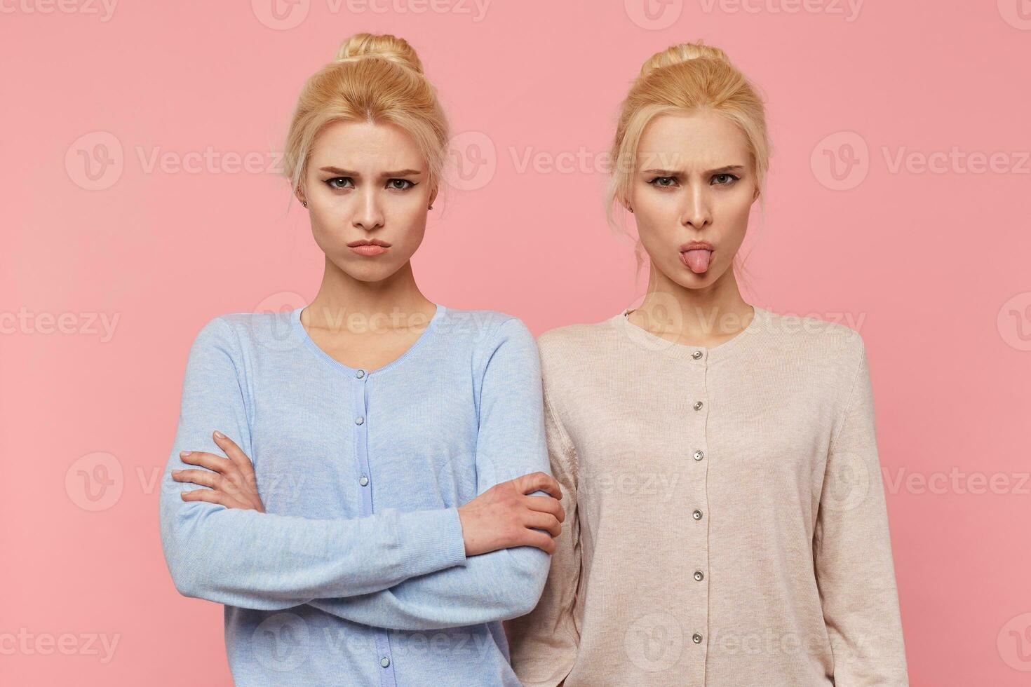 Frowning beautiful young blonde twins in a bad mood, looking in the camera isolated over pink background. photo