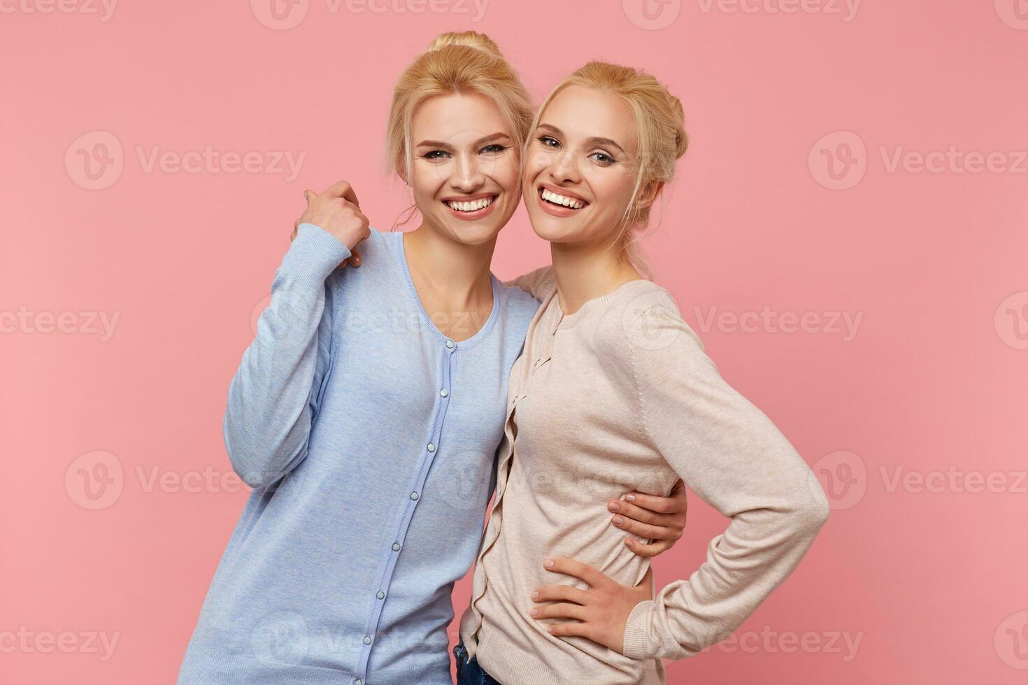 Photo of cute blonde twin sisters in identical cardigans of different colors, hugging each other, happy and funny, smiles broadly stads over pink background.