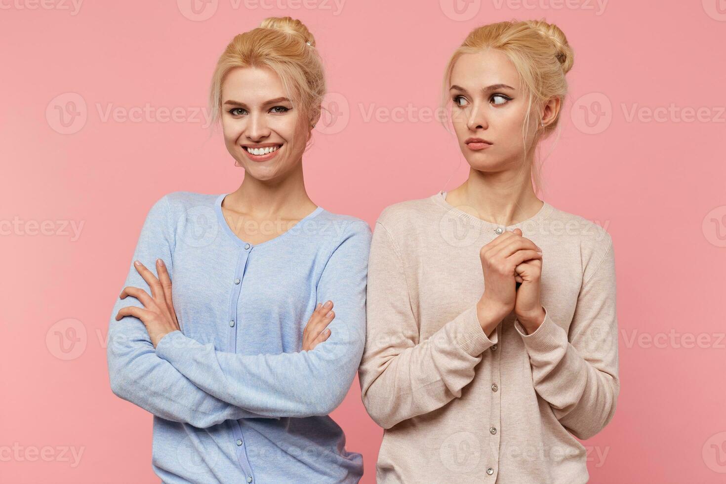 Young cute blonde woman frightened looks at her joyful sister isolated over pink background. photo