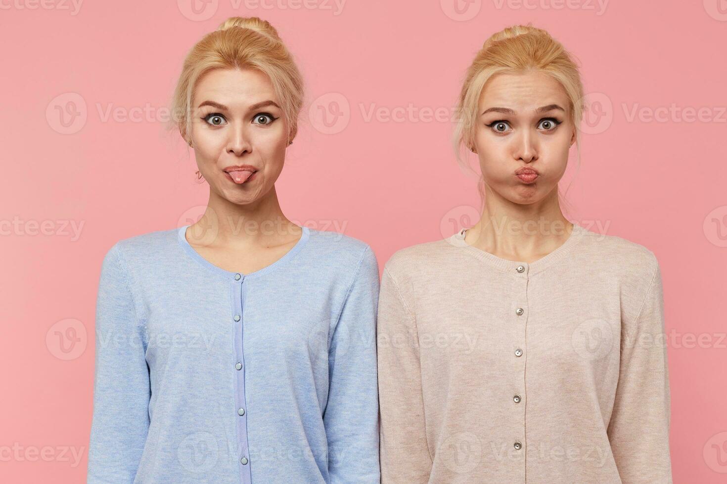 Photo of beautiful young blonde twins make faces, show tongue and blow cheeks, looking at the camera isolated over pink background.