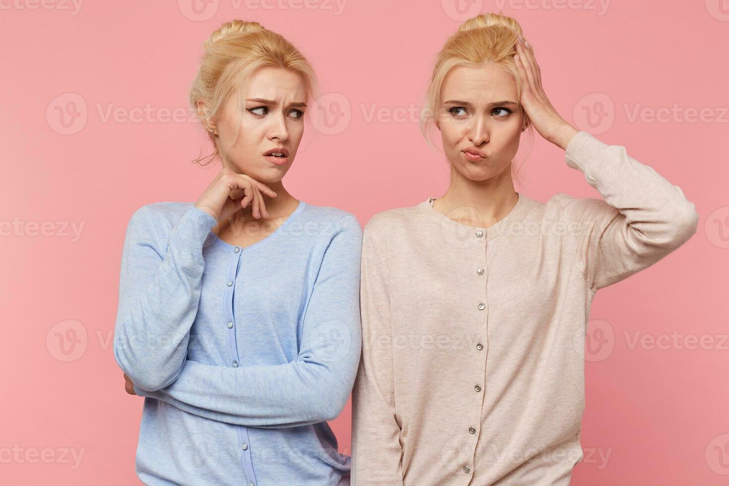 One of the beautiful young blonde twins forgot where the keys to the car, and her sister is perplexed and angry at her. Sisters isolated over pink background. photo