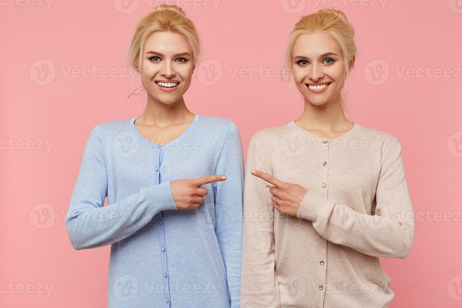 Photo of young broadly smiling happy blonde twins, pointing at each other isolated over pink background.