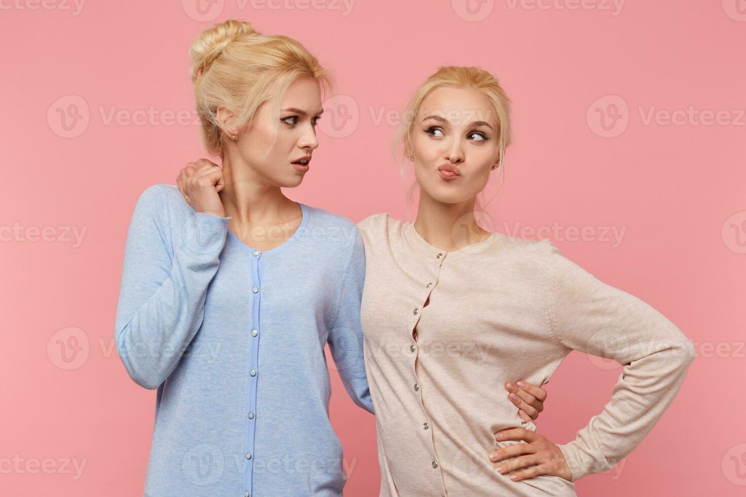 Portrait of cute twins blondes, one of the sisters is naughty and grimacing and the second is disapprovingly looking at her. Stands over pink background. People and emotions concept. photo