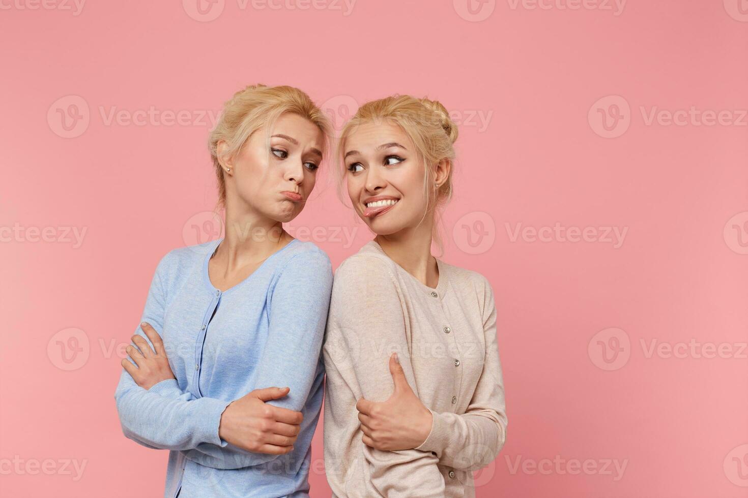 Portrait of cute twins blondes, girls fool standing with their backs to each other, grimace and and look at each other. Stands over pink background. photo