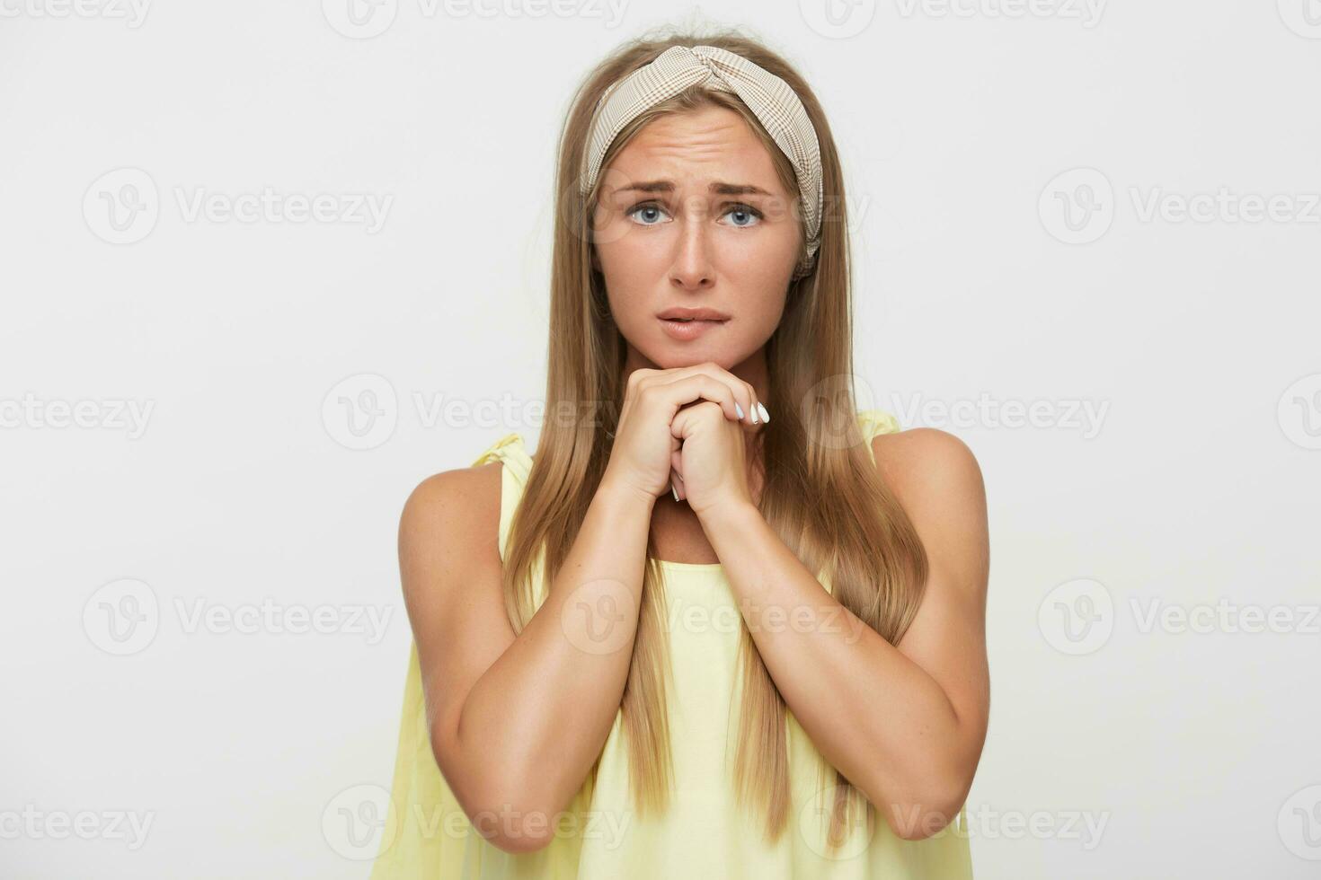 retrato de joven nervioso atractivo rubia hembra ceñudo su Cejas y mordiendo preocupantemente debajo del labio mientras posando terminado blanco fondo, plegable elevado manos debajo su barbilla foto