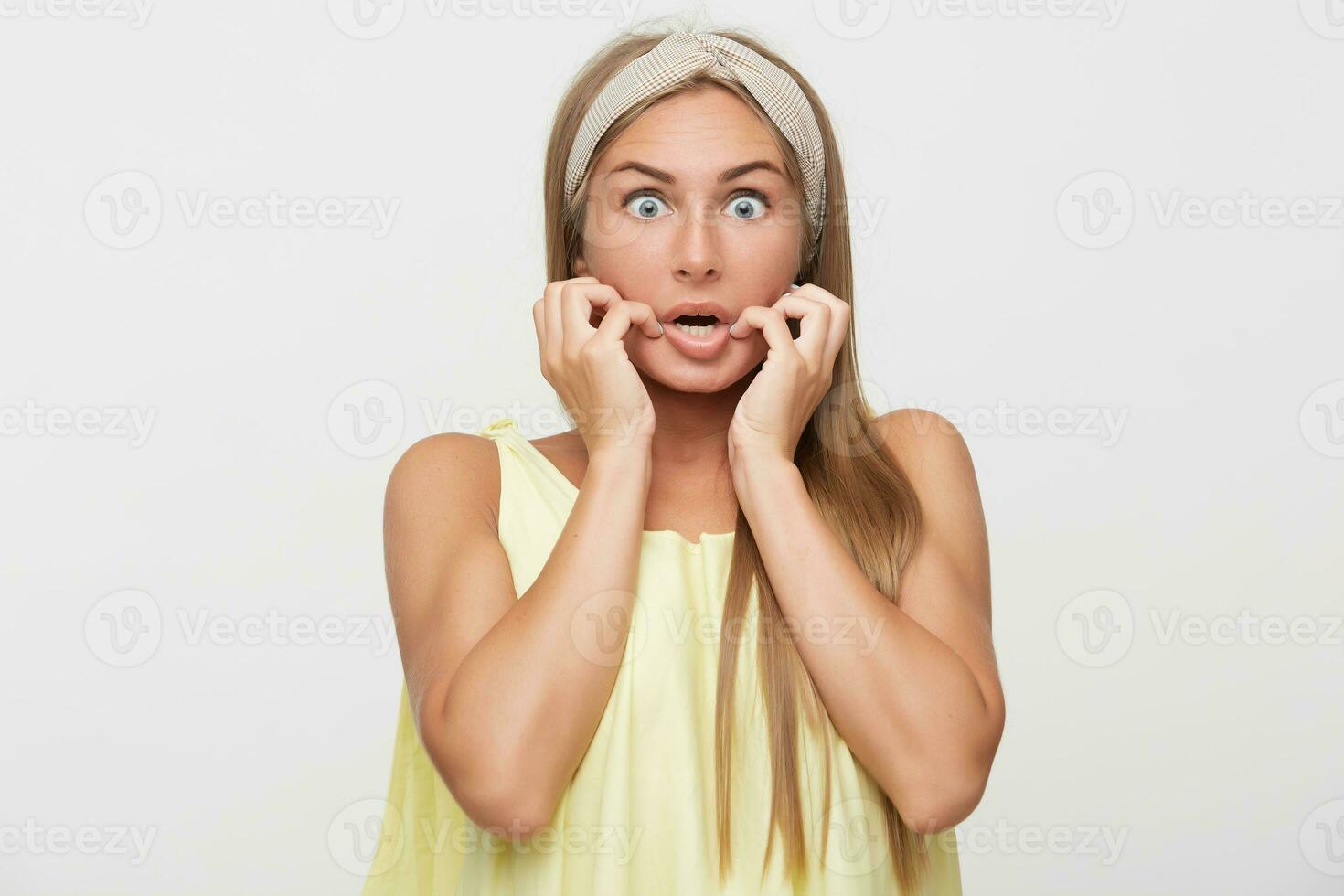 Shocked young long haired blonde female in beige headband raising scaredly hands to her face and rounding her blue eyes while looking amazedly at camera, isolated over white background photo