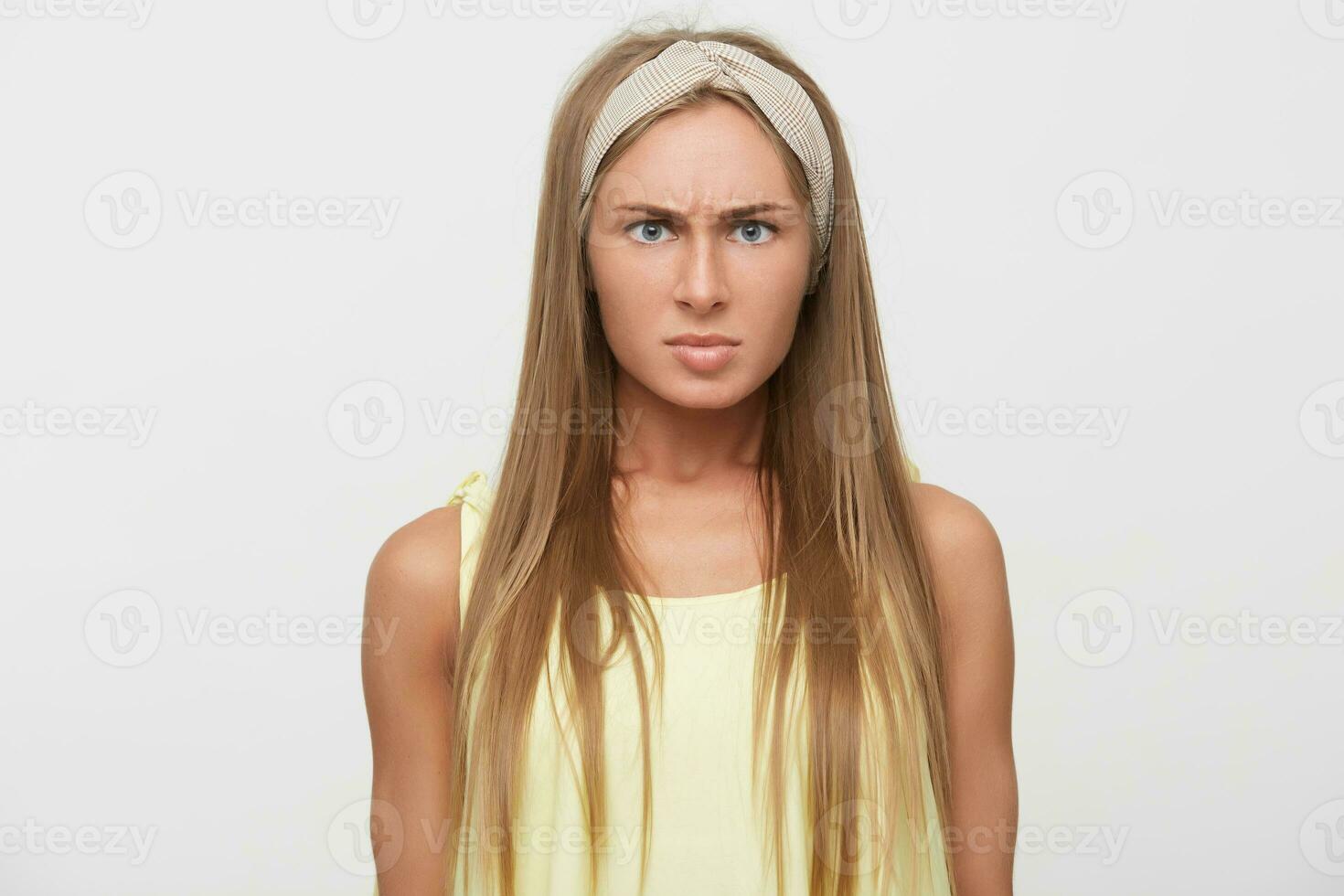 Scowling young beautiful blonde female keeping her lips folded while looking scowlingly to camera, dressed in casual clothes while posing over white background with hands down photo